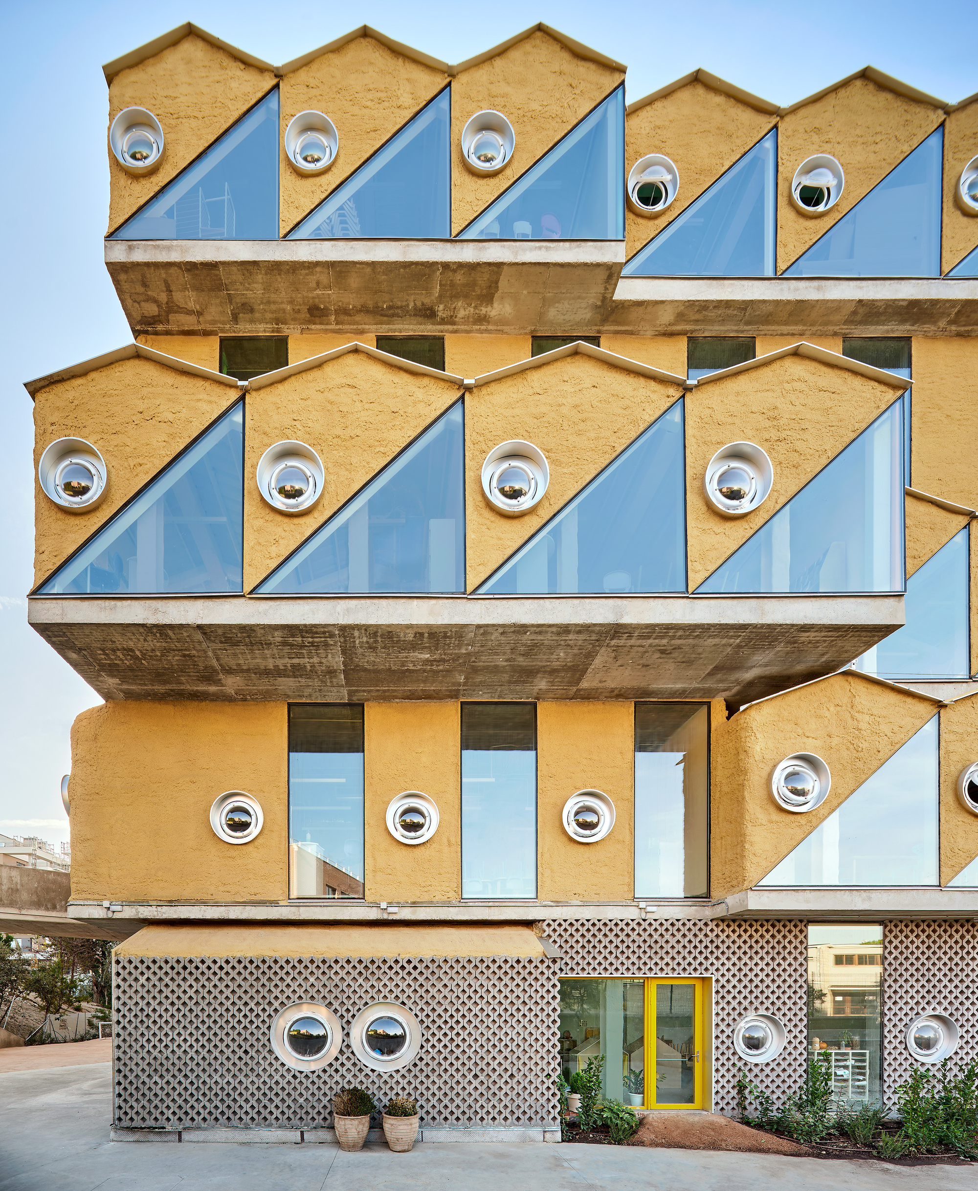 Stacked concrete floors with triangular blue panels and circular porthole windows under a clear sky