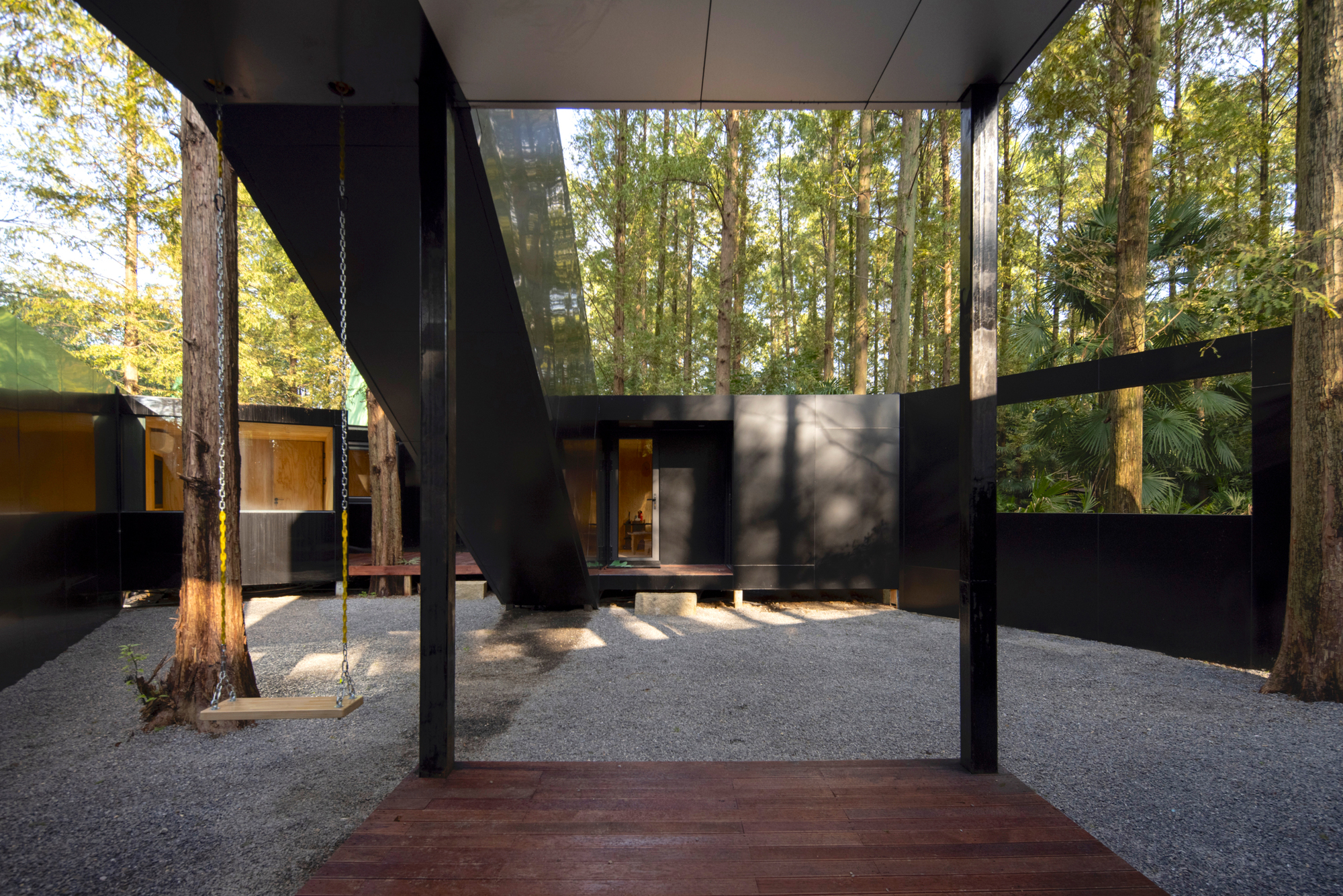 View from the timber deck through the courtyard gate toward the black steel volumes and preserved eucalyptus trunks