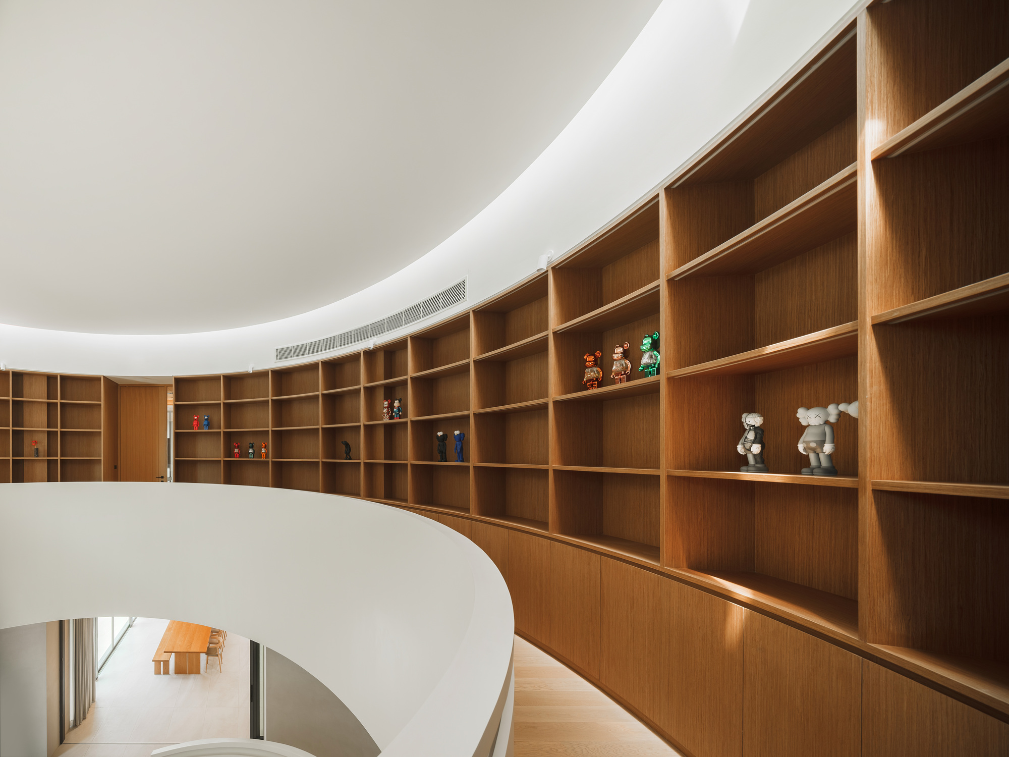 Curved oak shelving wall with collectible toys displayed above the white spiral stair void