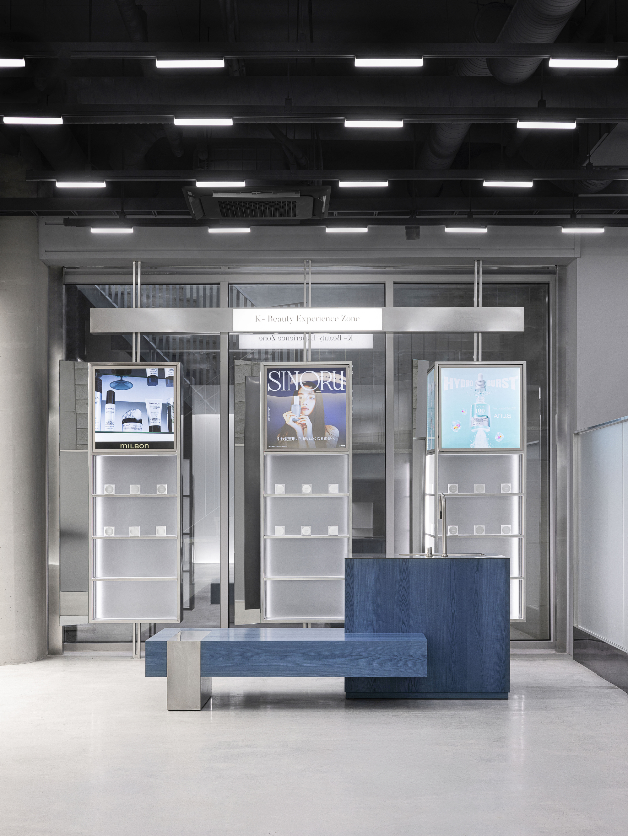 Glass entrance with product displays and blue bench under grid of linear ceiling fixtures