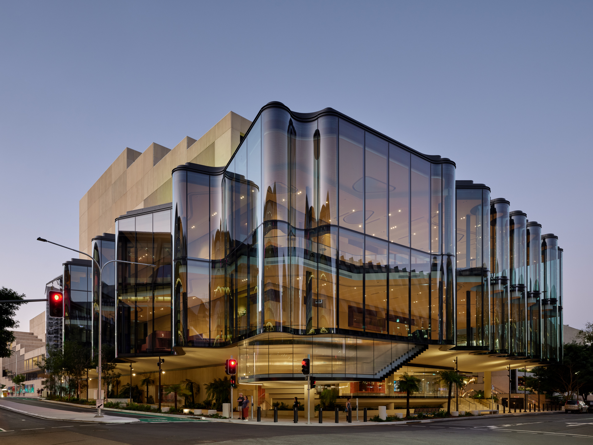 Street corner view of the undulating glass facade with vertical cylindrical tubes lit at dusk