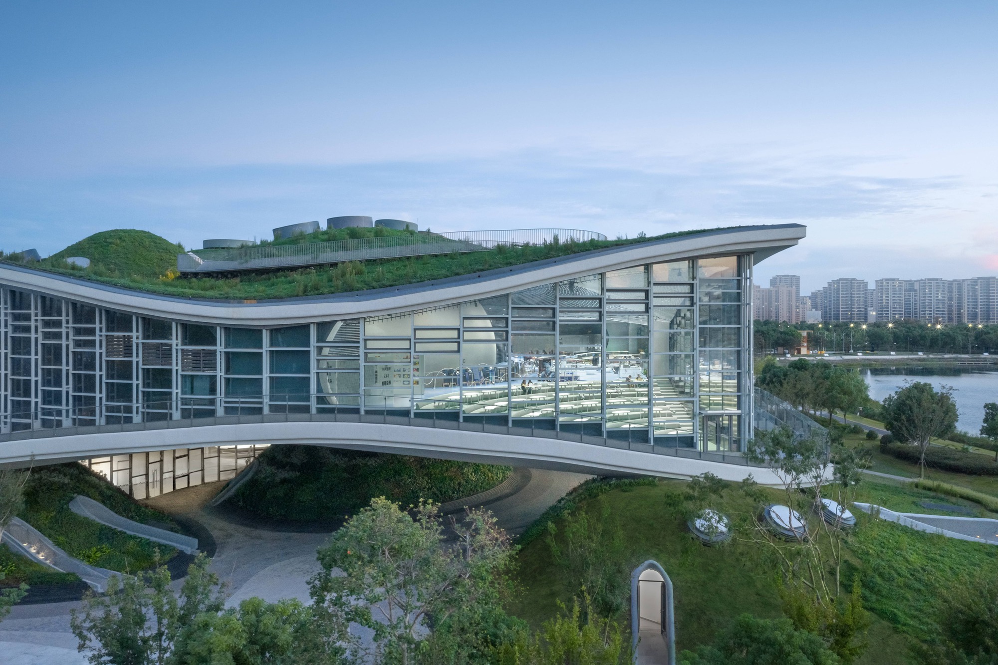 Glass curtain wall and cantilevered roof edge at dusk with planted slopes descending to the ground level