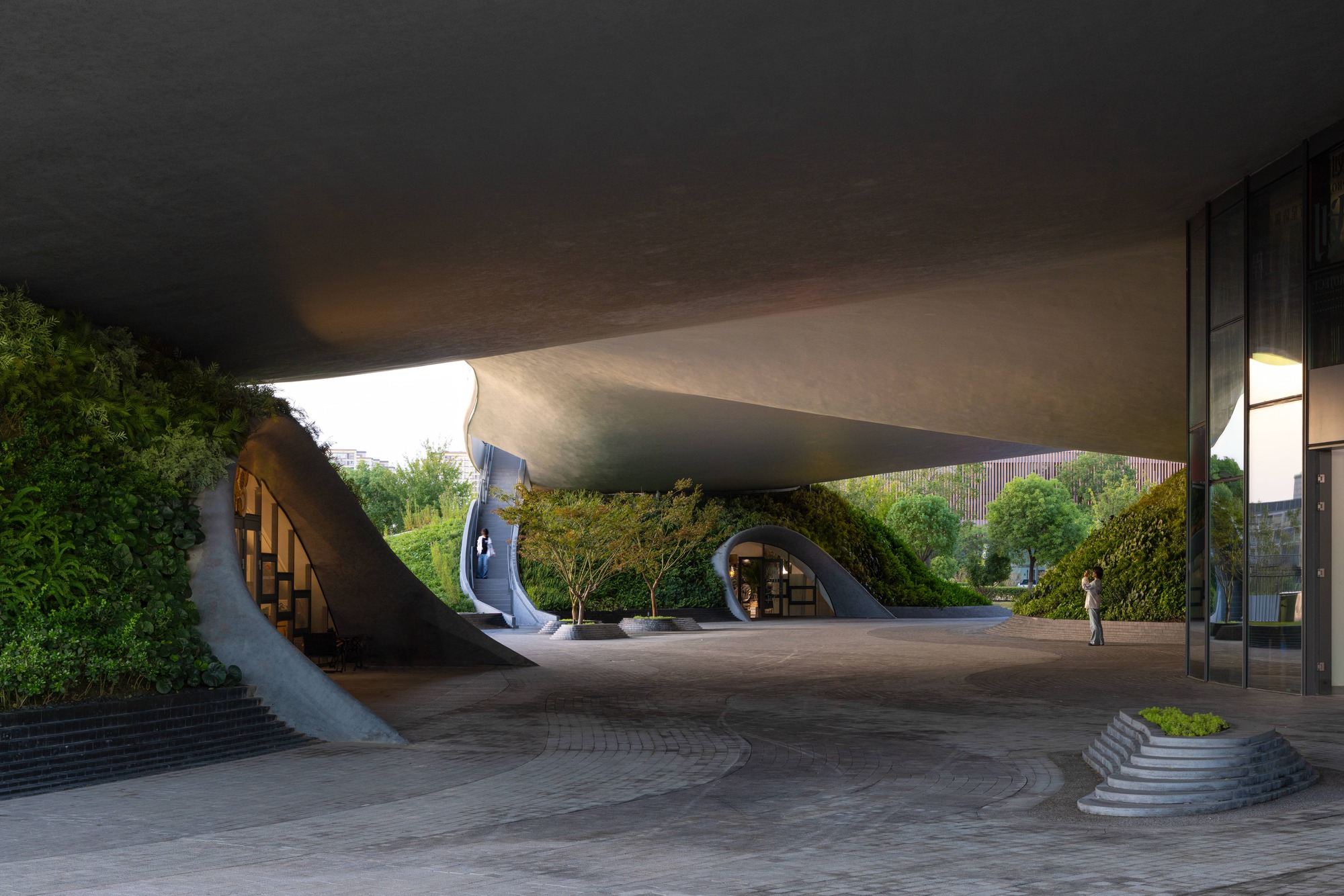Covered forecourt beneath the concrete shell roof with curved stairs ascending to the planted roof