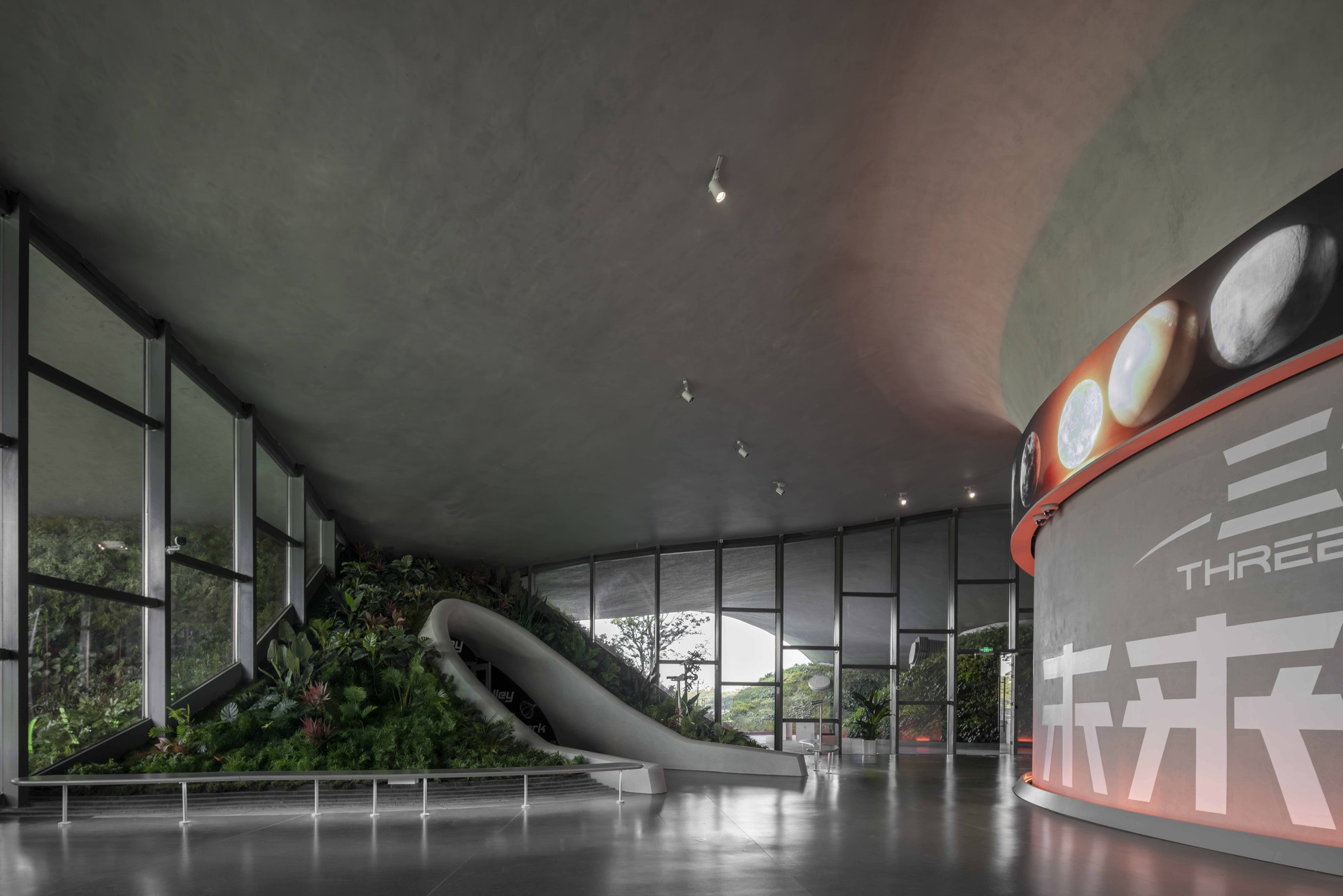 Interior space with floor-to-ceiling glazing, planted hillocks, and a tubular slide under concrete ceiling