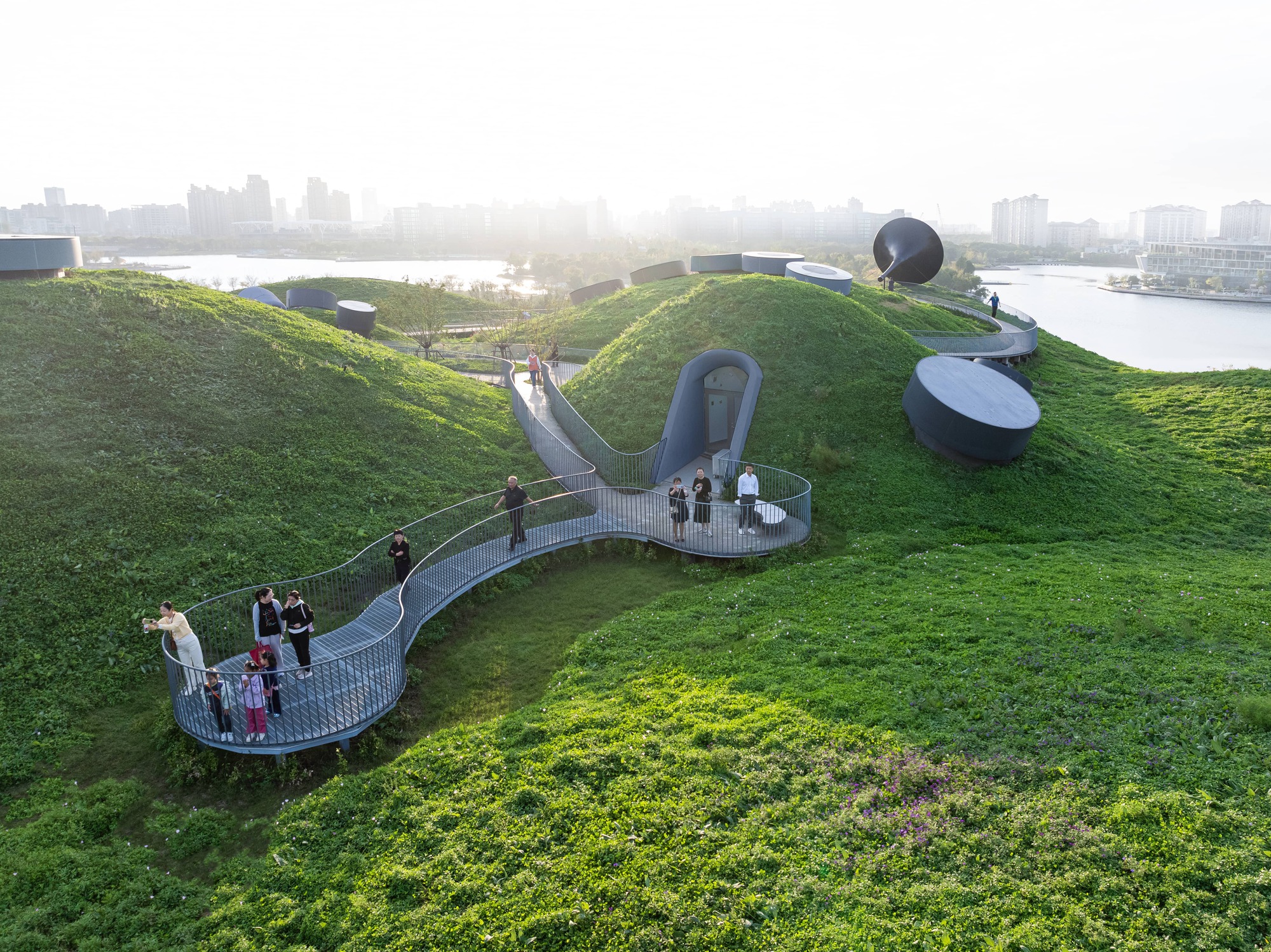 Steel mesh walkway winding across the planted green roof with visitors gathered on viewing platforms at dusk
