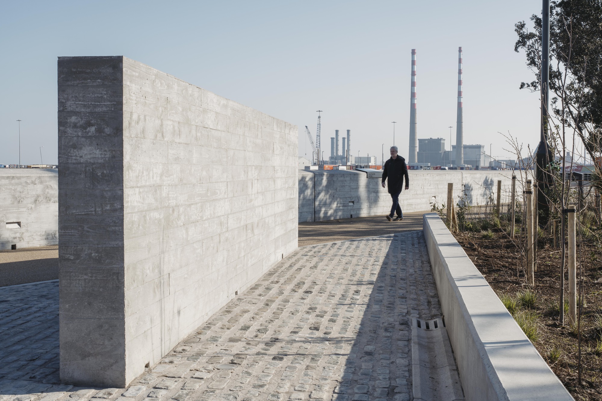 Cobblestone pathway along concrete parapet wall with pedestrian and industrial stacks in distance