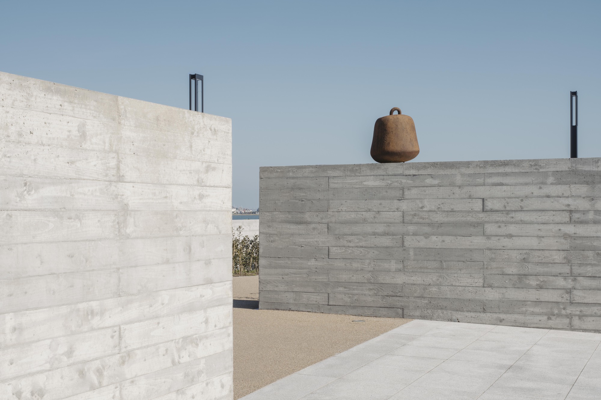 Courtyard framed by board-formed concrete walls with weathered bell suspended from upper parapet