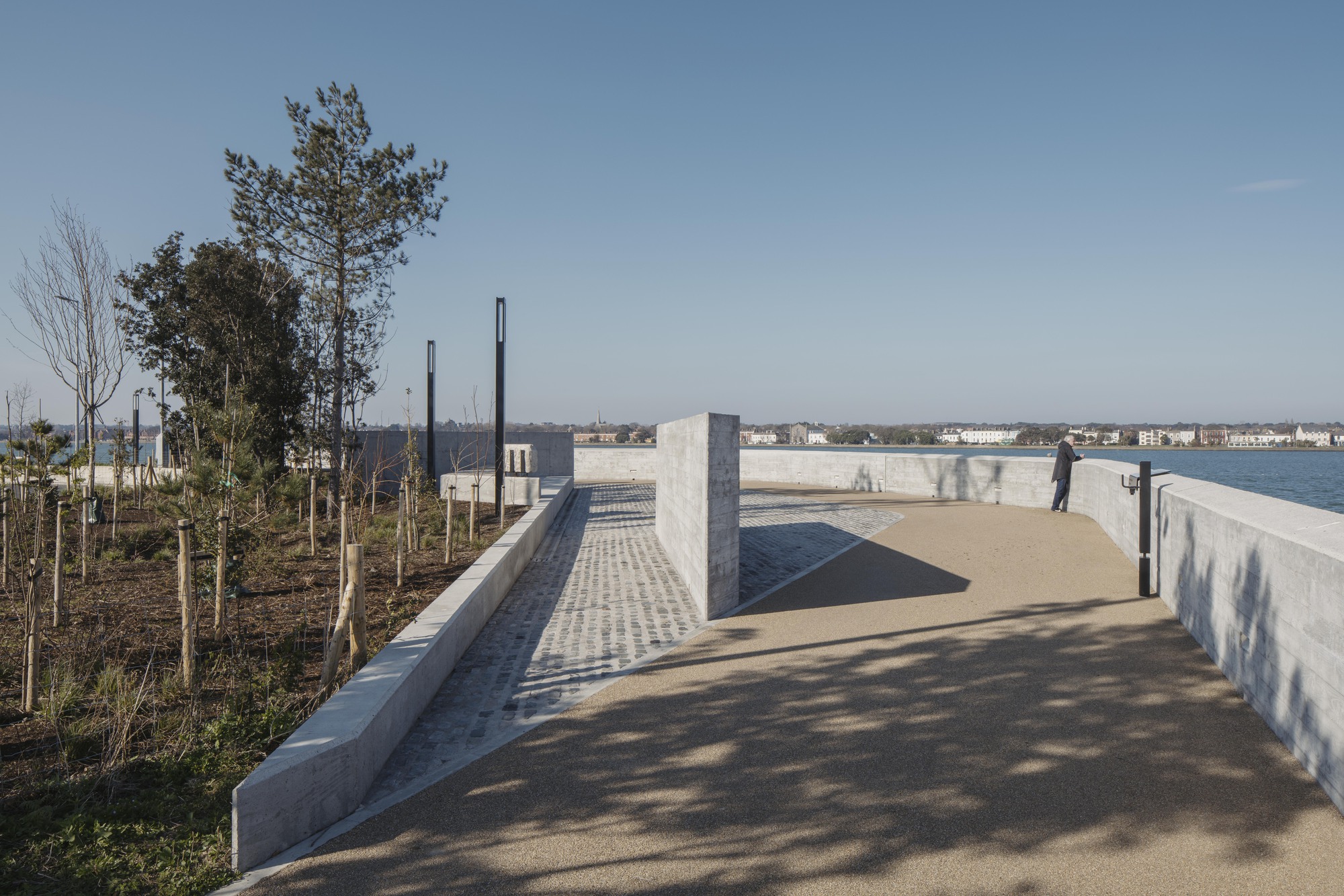 Curving pathway bordered by concrete wall and planted beds with young trees overlooking the bay