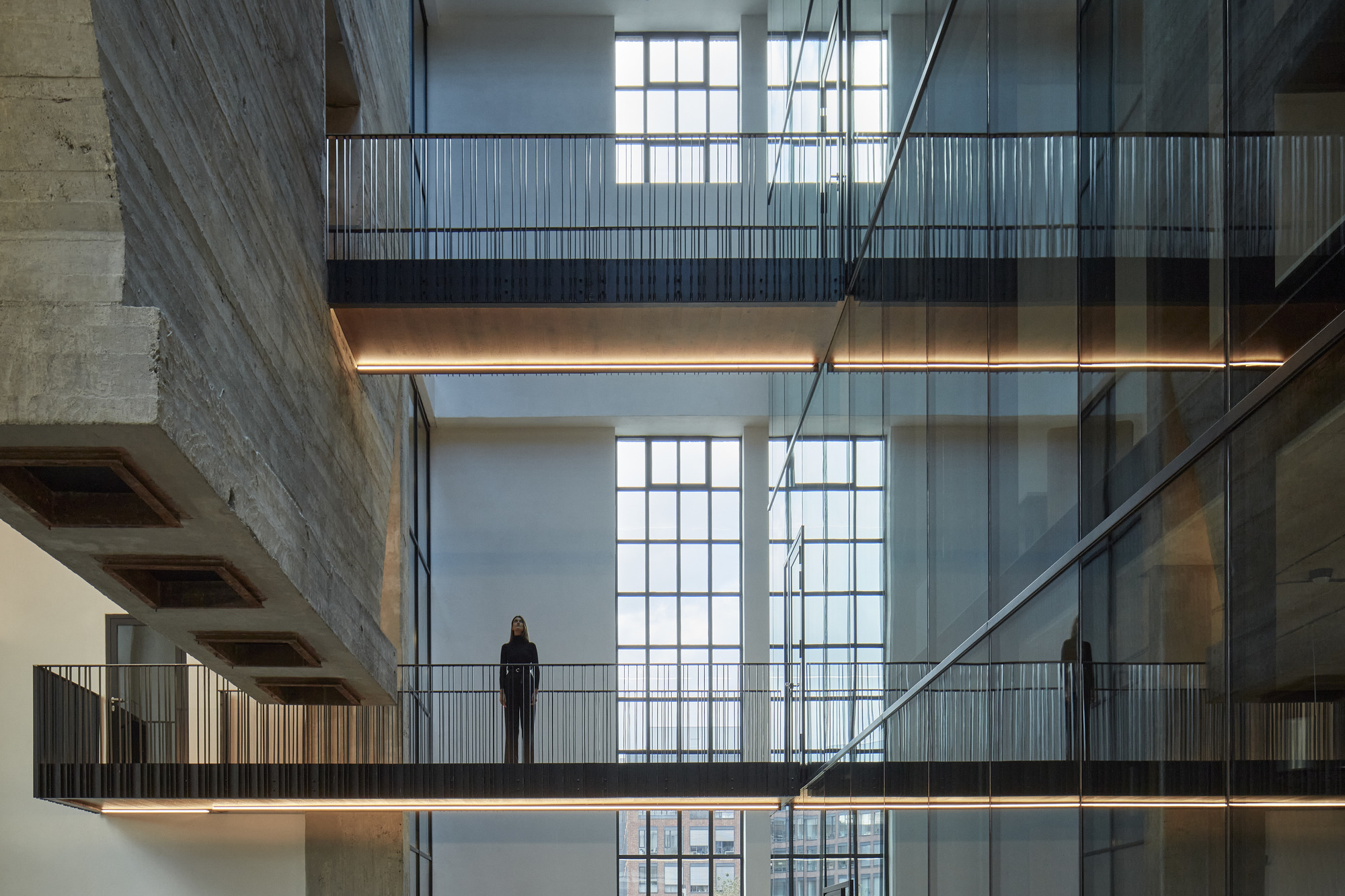 Multilevel atrium with suspended glass bridges, ribbed metal railings, and a figure standing against gridded windows