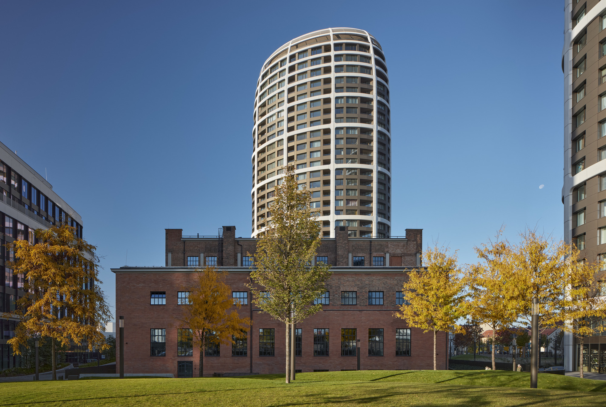 Cylindrical tower rising above a retained brick warehouse with trees in autumn foliage on a lawn