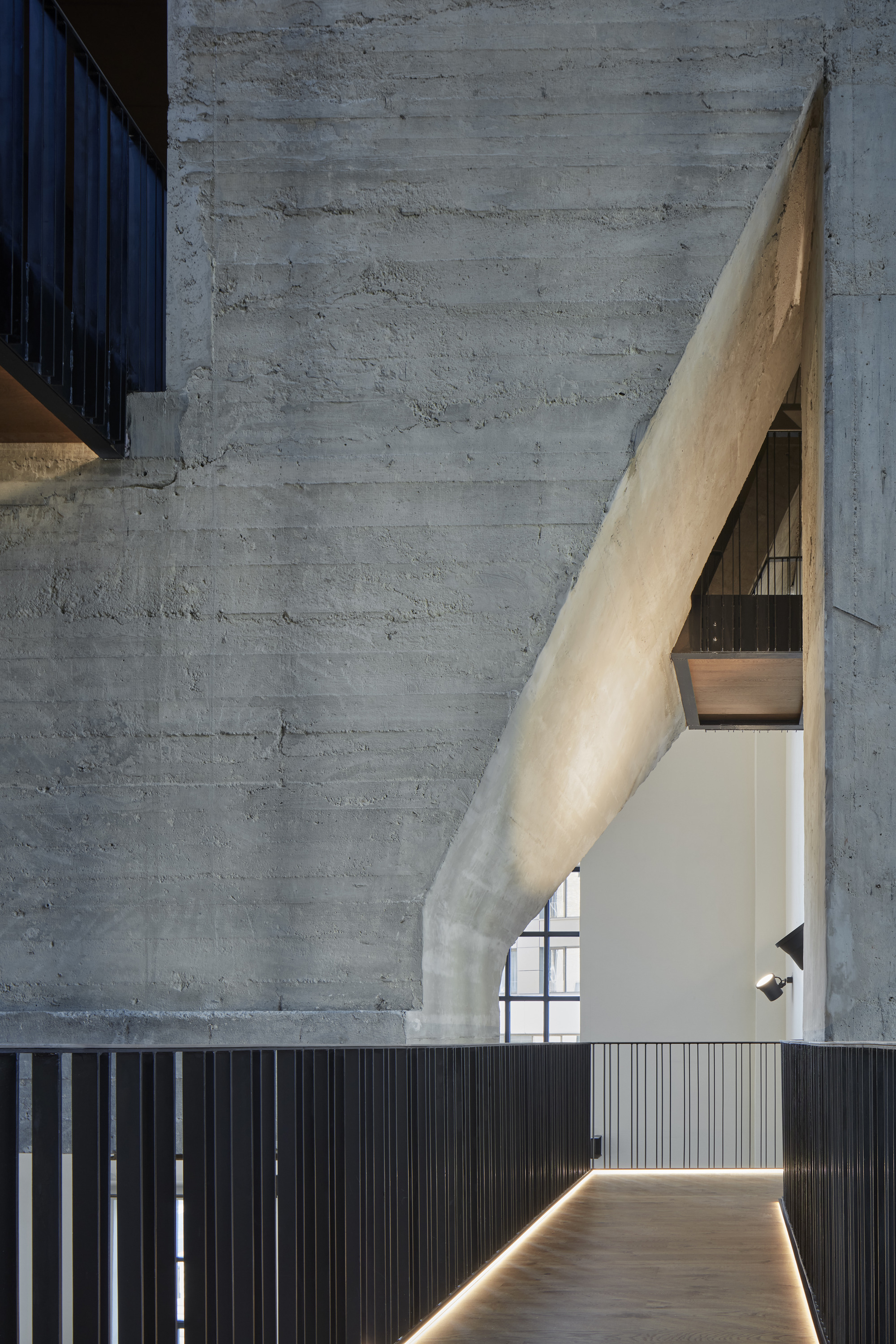 Triangular concrete beam leaning against board-formed walls above a backlit corridor with vertical metal cladding