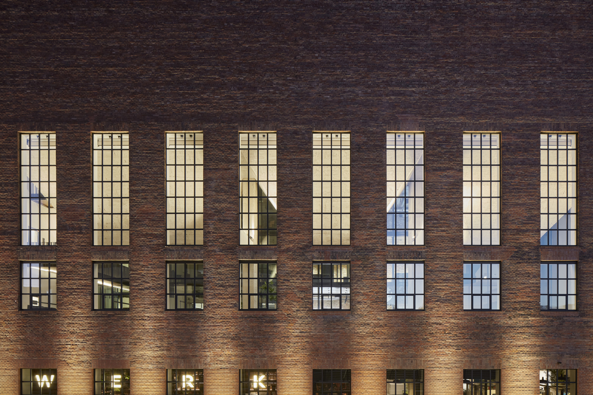 Dark brick facade with tall gridded windows illuminated from within at dusk