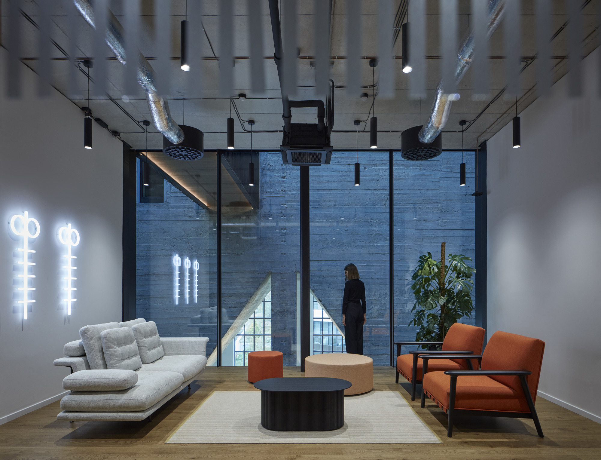 Lounge area with orange chairs, white sofa and figure standing at tall windows overlooking brick wall