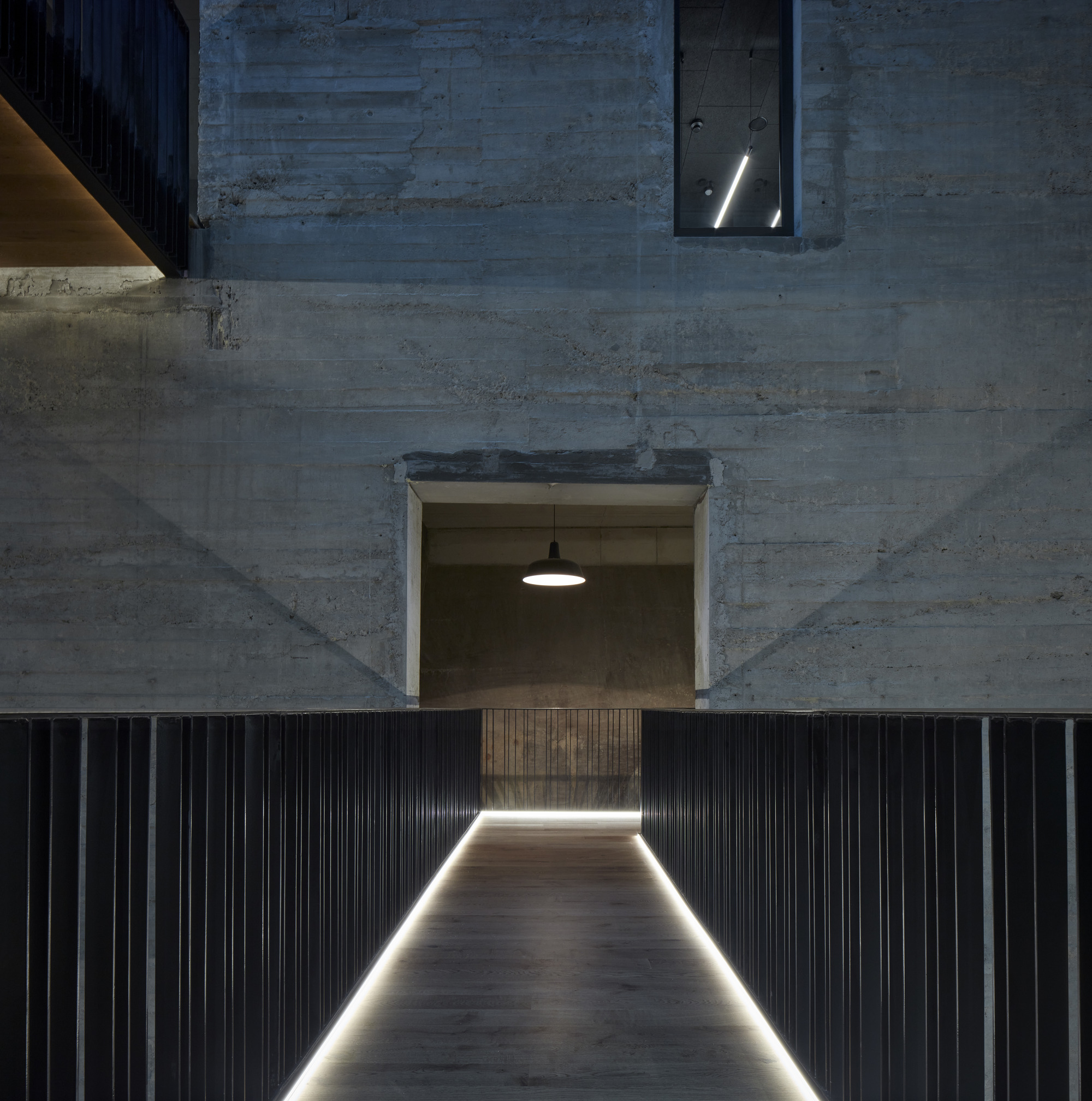 Board-formed concrete wall with window opening above corrugated metal panel base with recessed floor lighting