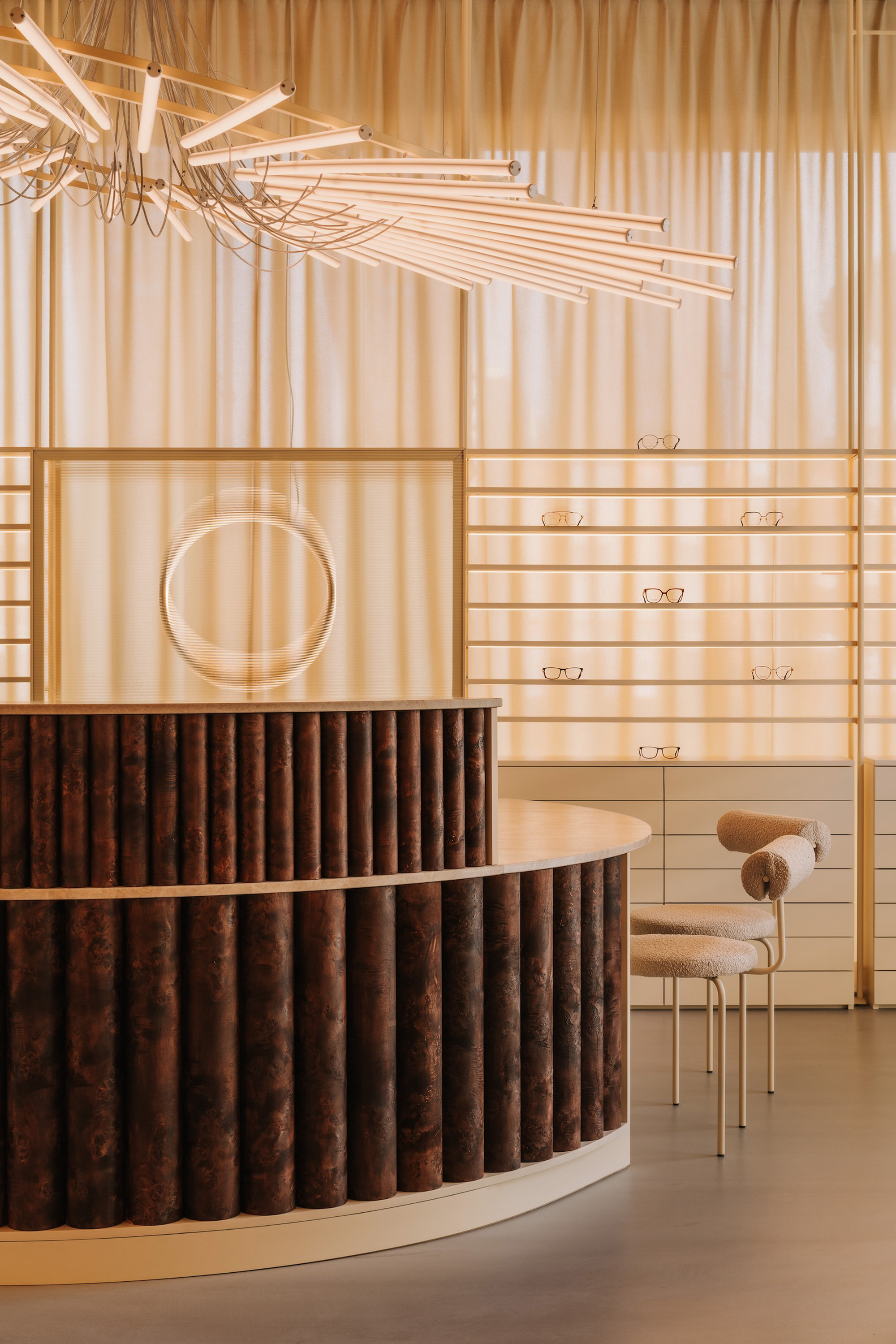 Curved reception desk clad in vertical timber slats beneath a radiating tubular light fixture and translucent curtain