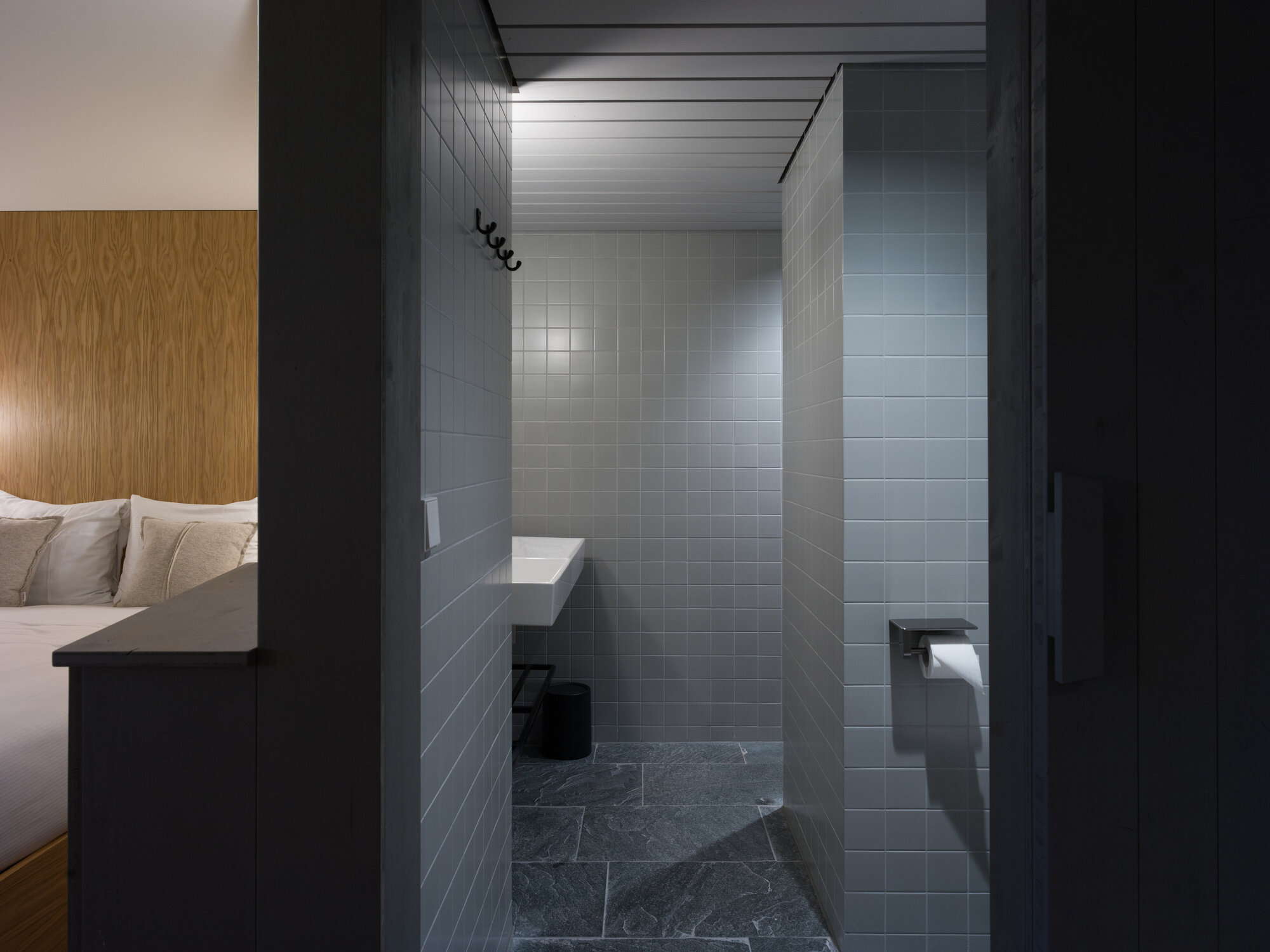 White-tiled shower enclosure with black fixtures viewed through a dark doorway from the bedroom