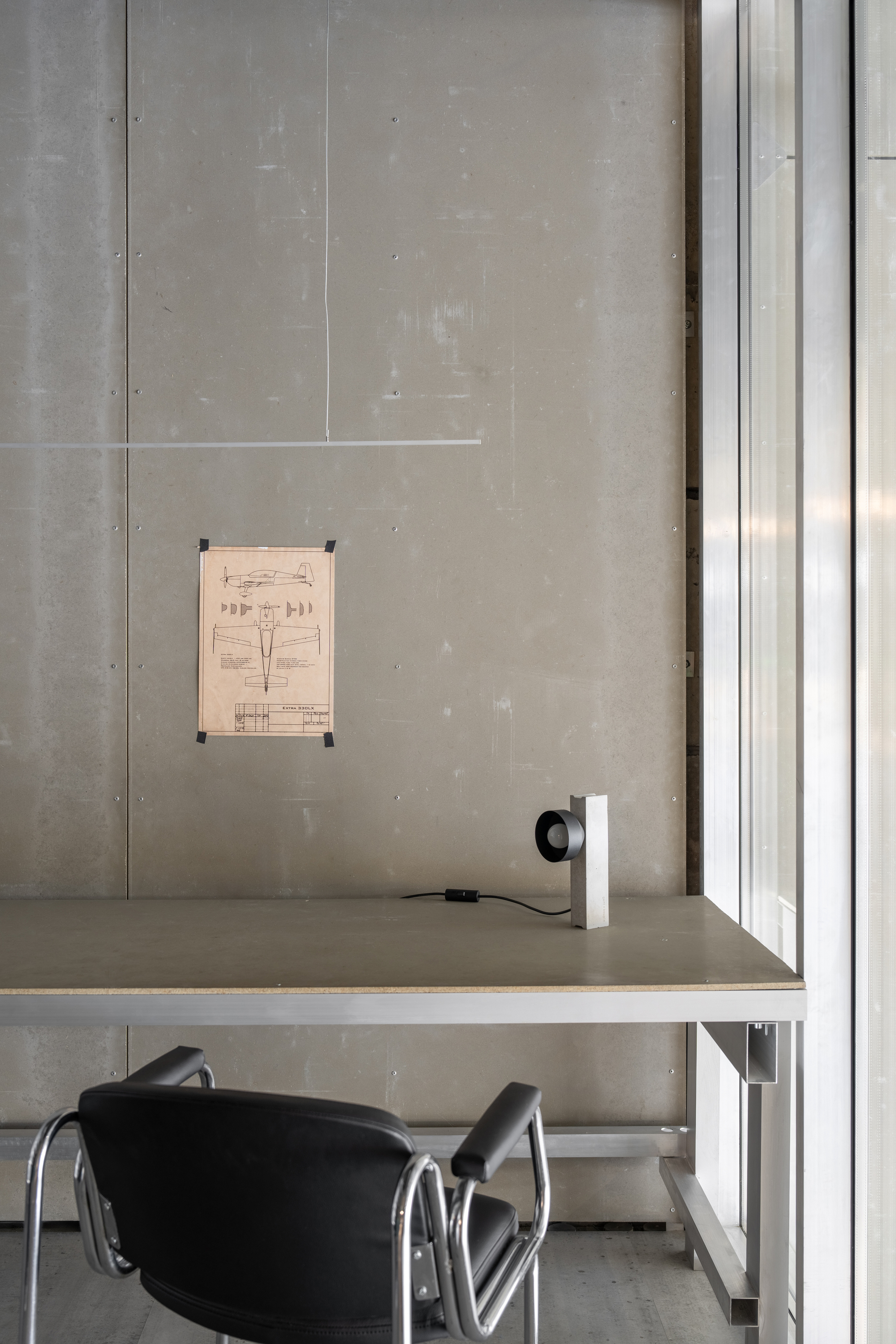 Workspace with concrete wall, window detail, suspended technical drawing, and black chair at desk