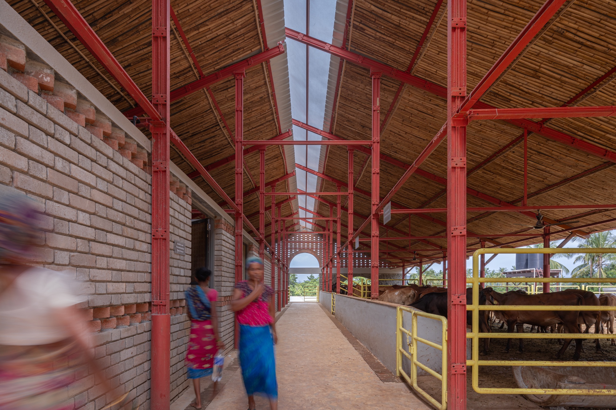 Covered walkway with red steel frame and woven bamboo ceiling alongside a brick wall and cattle enclosure