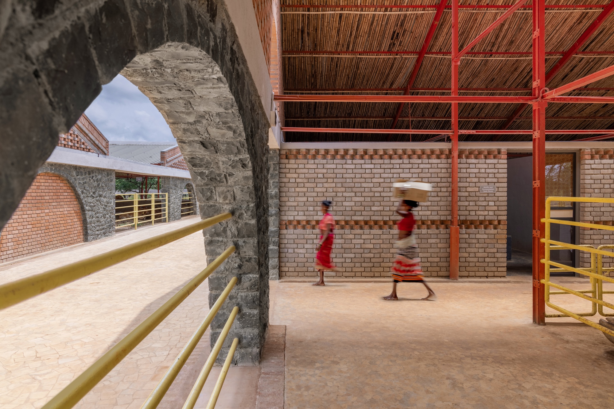 Stone arch opening framing a walkway with yellow railings and two figures passing beneath the bamboo roof