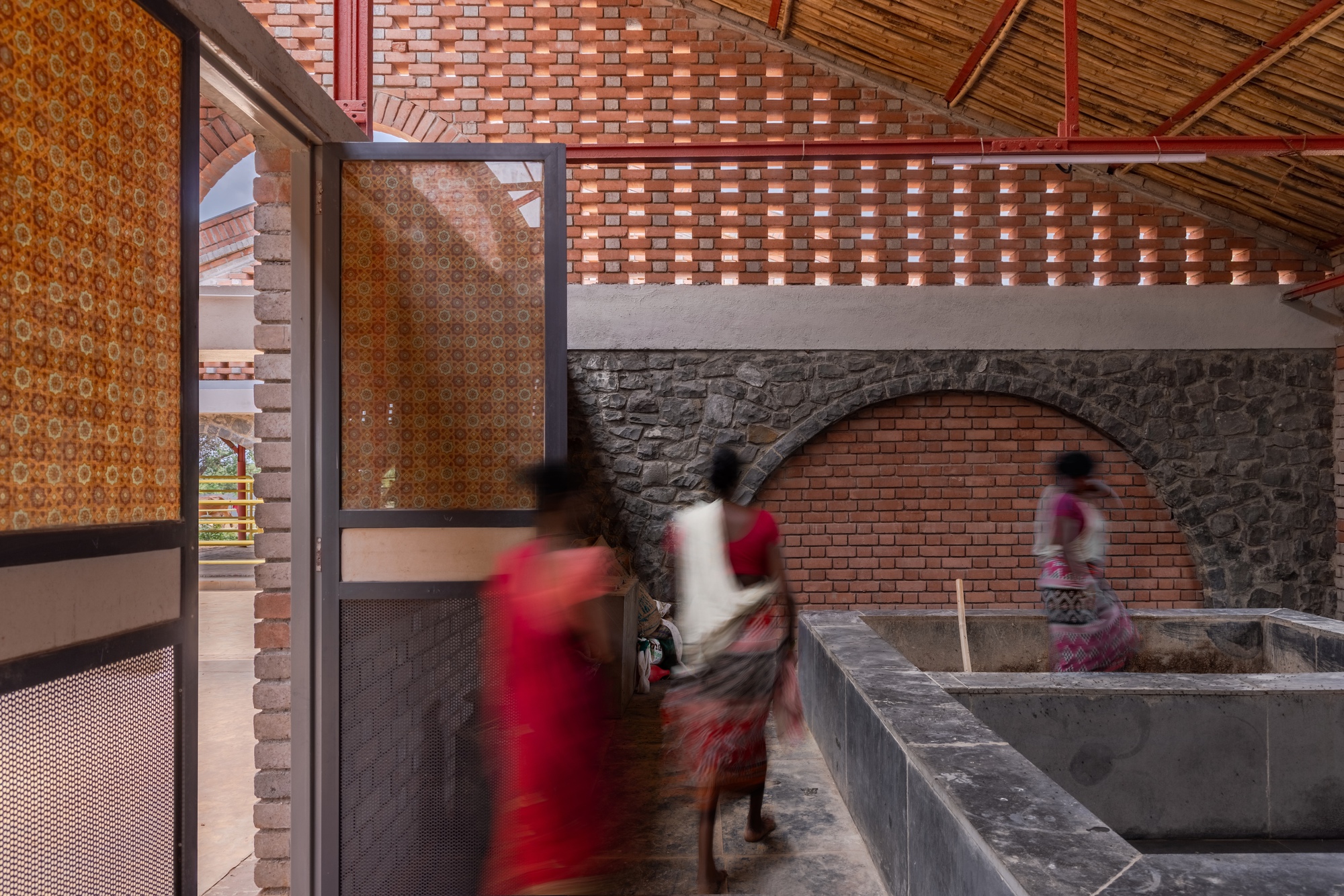 Interior corridor with perforated brick screen walls and concrete water troughs where figures move through filtered light