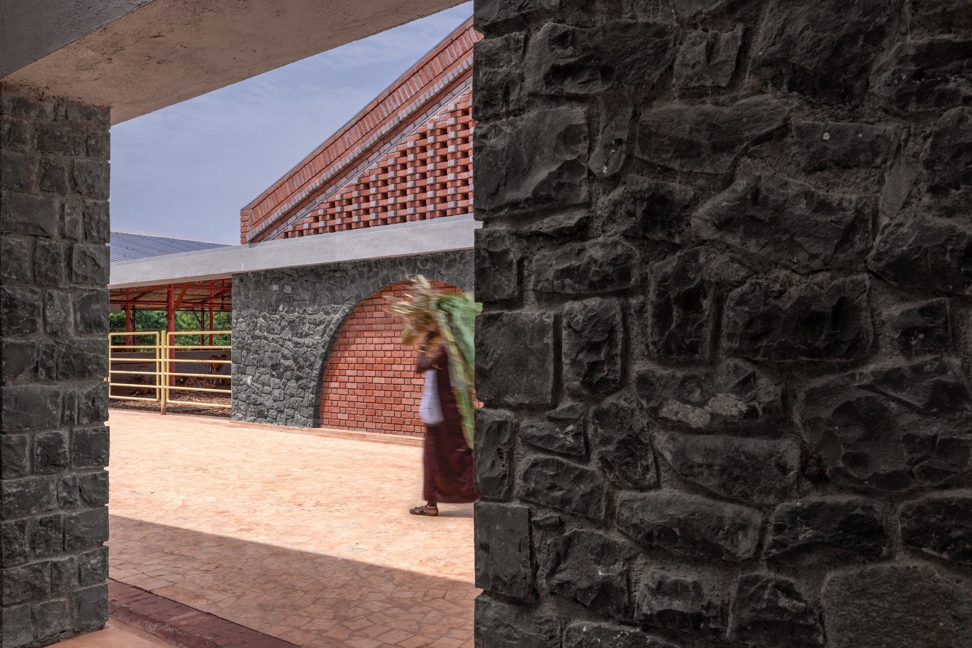 View through basalt stone columns showing a person walking across the courtyard toward the arched brick walls