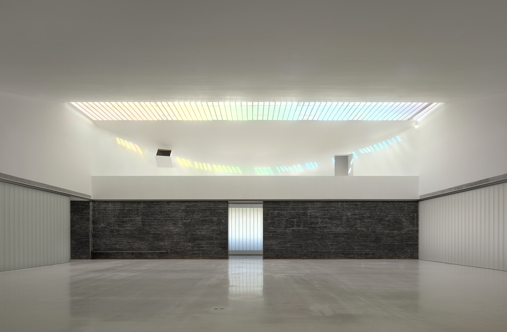 Interior hall with slatted skylight casting colored light onto a dark stone wall and polished floor
