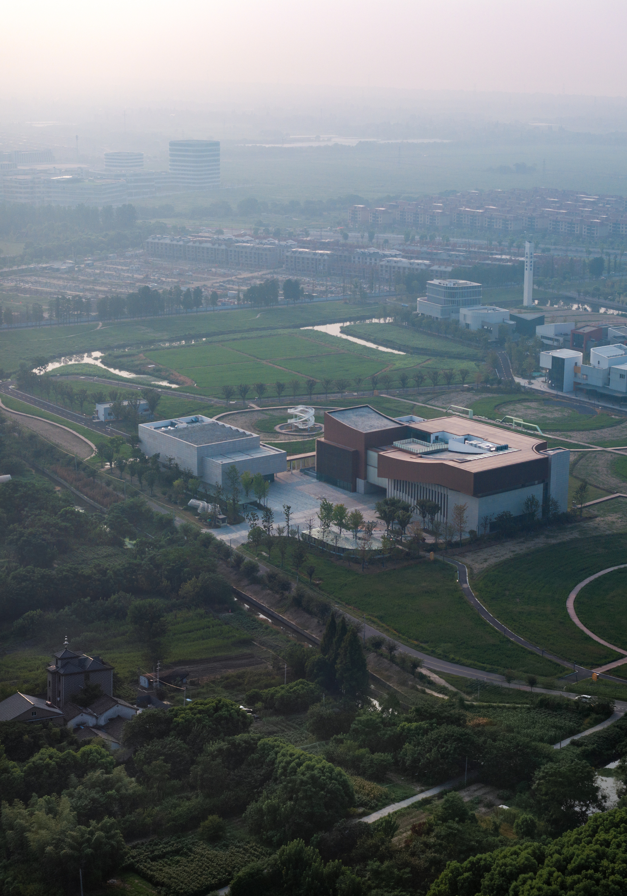 Aerial view of two connected volumes surrounded by green fields and agricultural landscape in morning haze