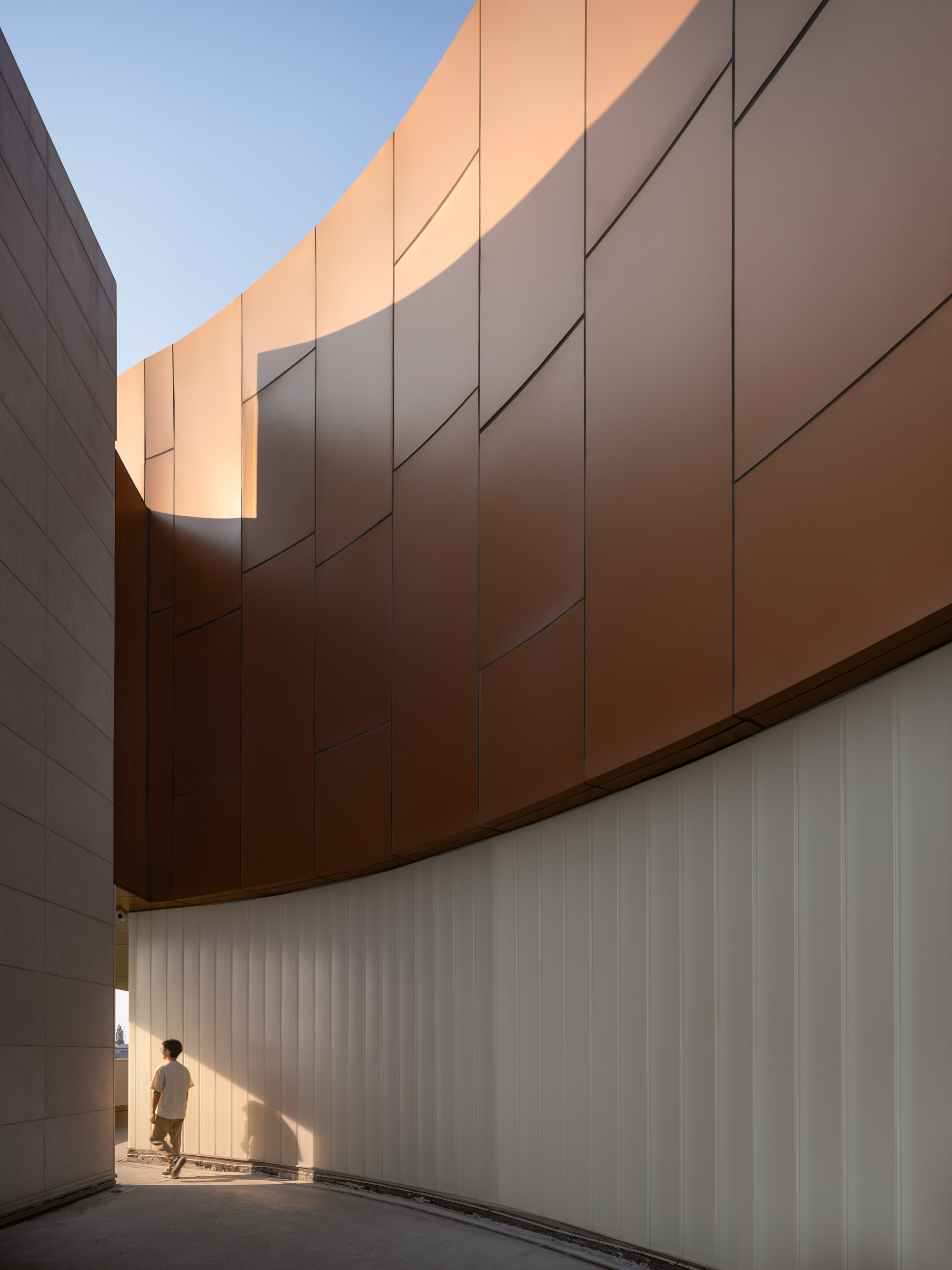 Courtyard passage with copper cladding above white vertical fins and a figure walking through afternoon light