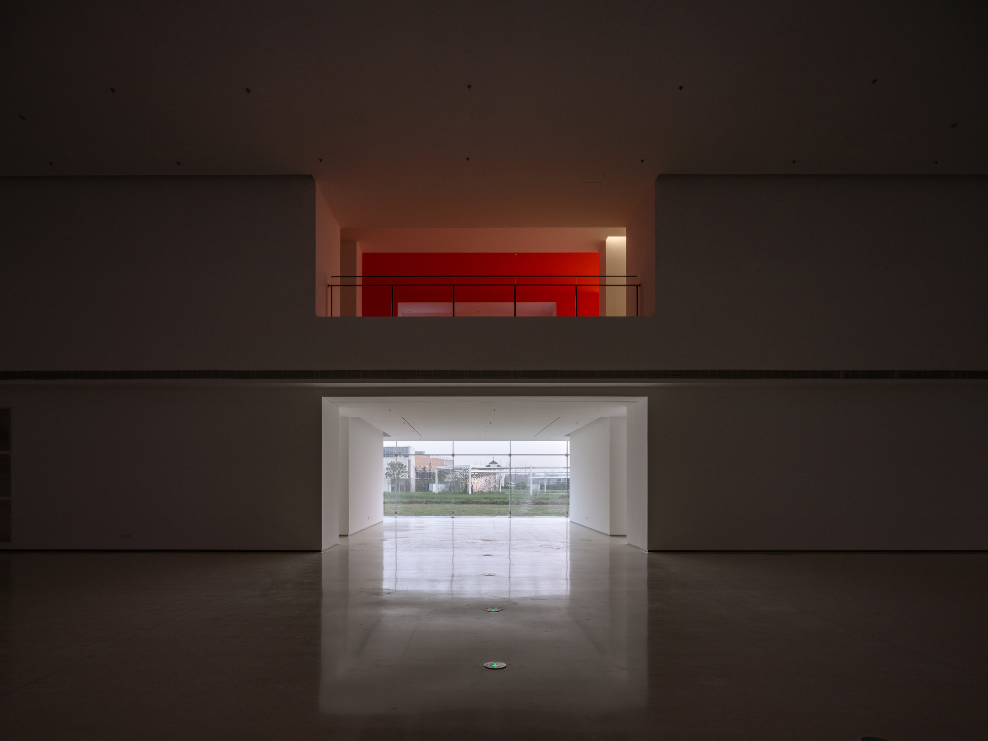 View through double-height void showing red-lit upper gallery and ground-level glazed entrance to landscape beyond