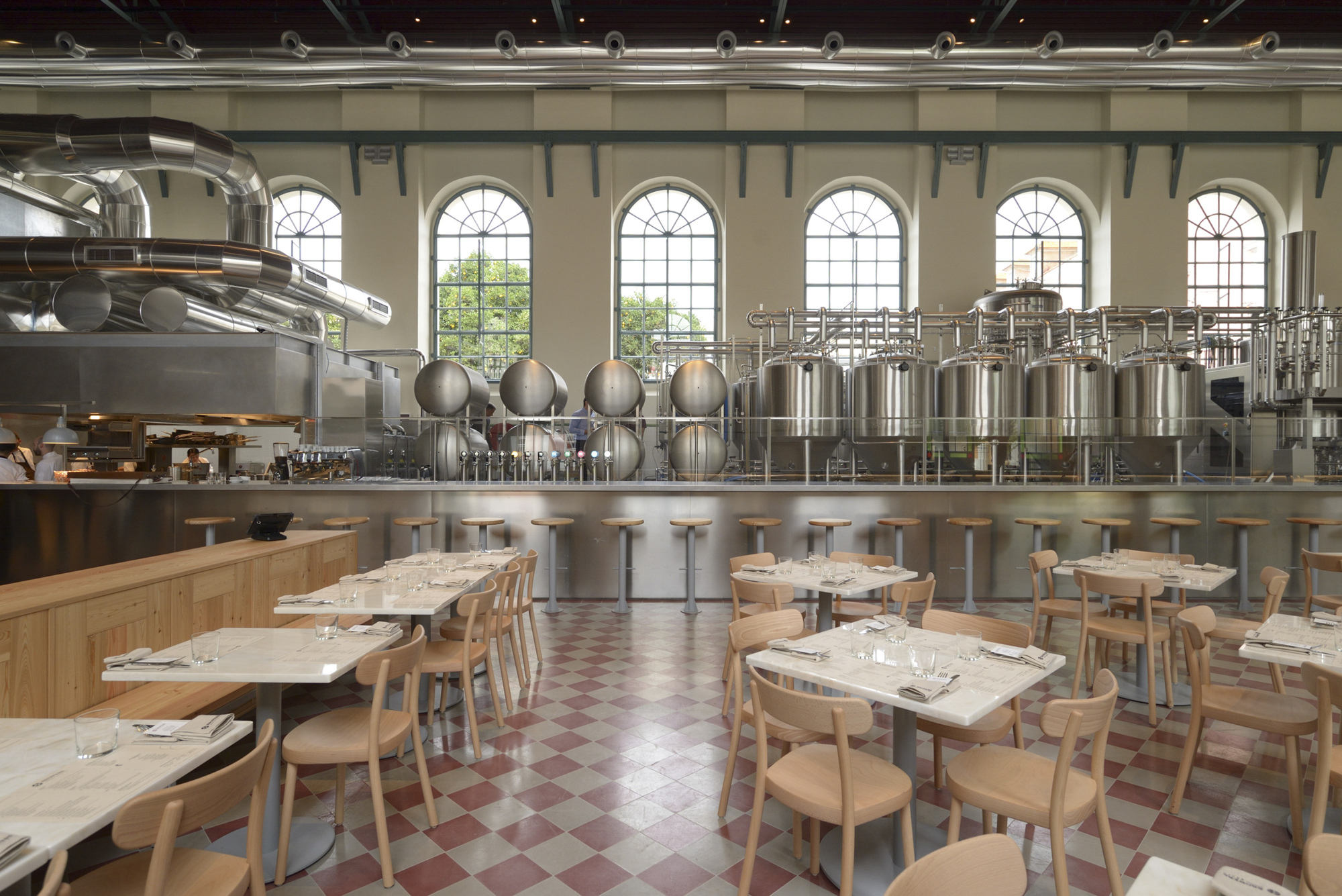 Dining area with timber tables and chairs on chequered flooring beside exposed stainless steel brewing tanks