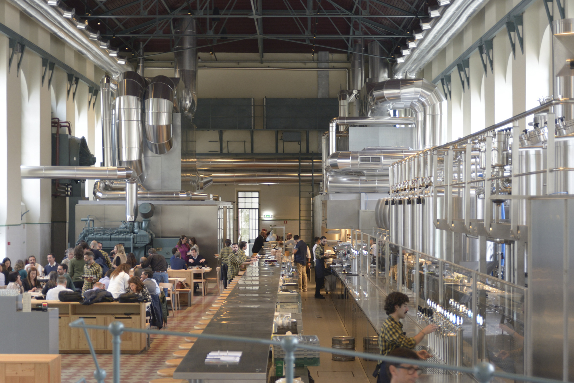 Hall filled with diners at timber tables surrounded by elevated stainless steel tanks and industrial piping