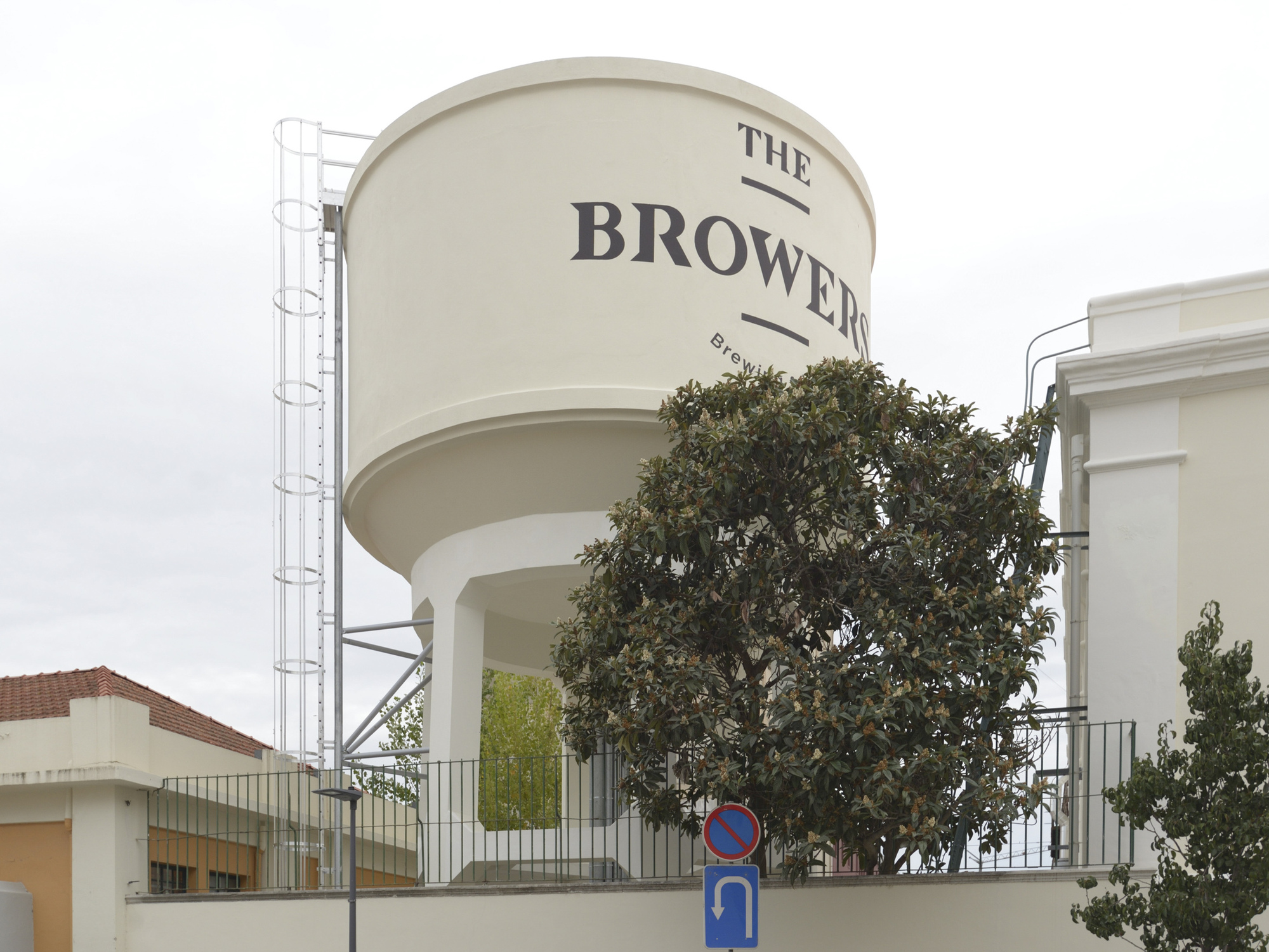 Cylindrical water tower with signage partially obscured by a mature tree in the foreground