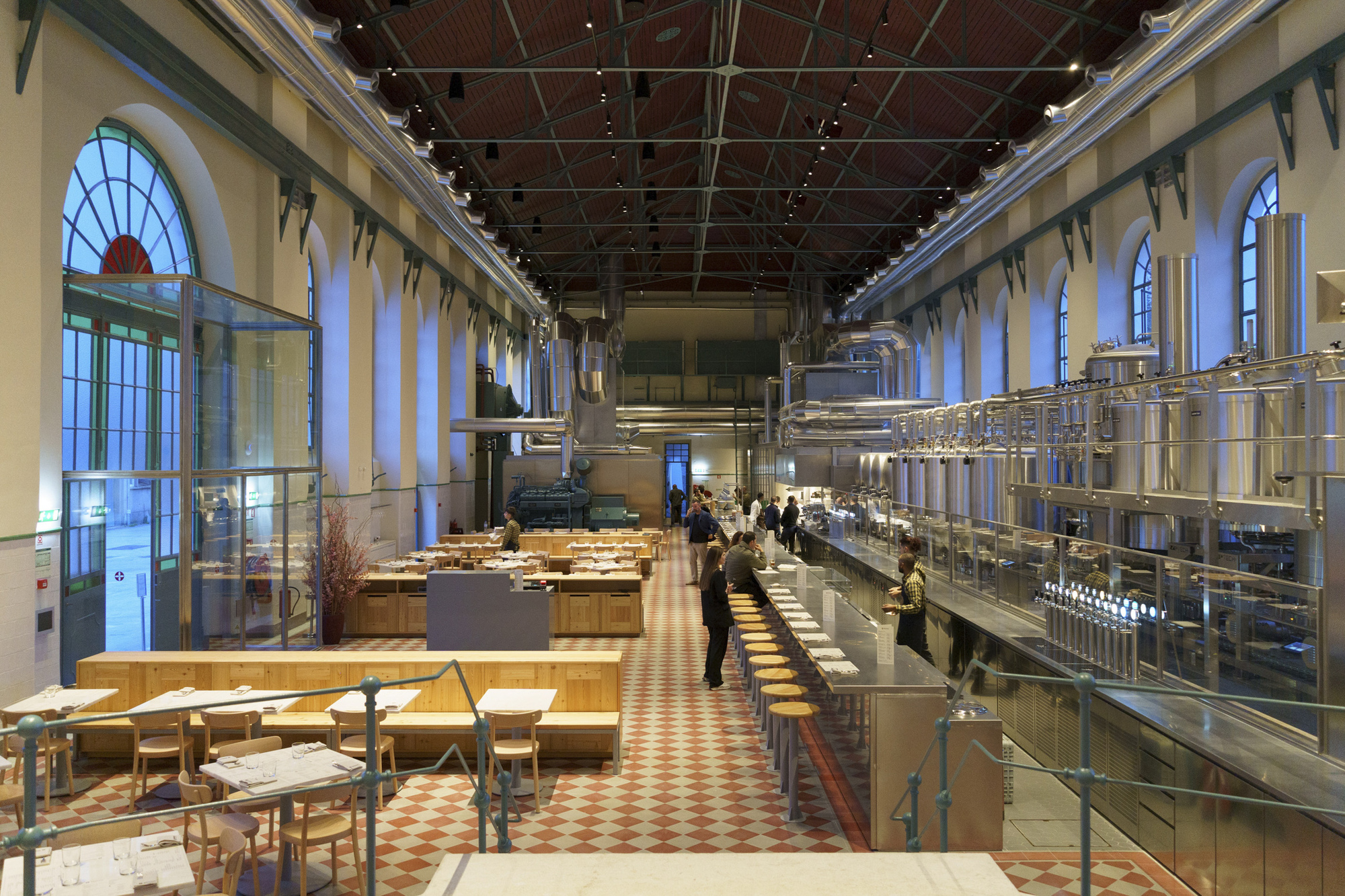 Long view of the hall showing exposed steel roof structure above timber tables and open kitchen area
