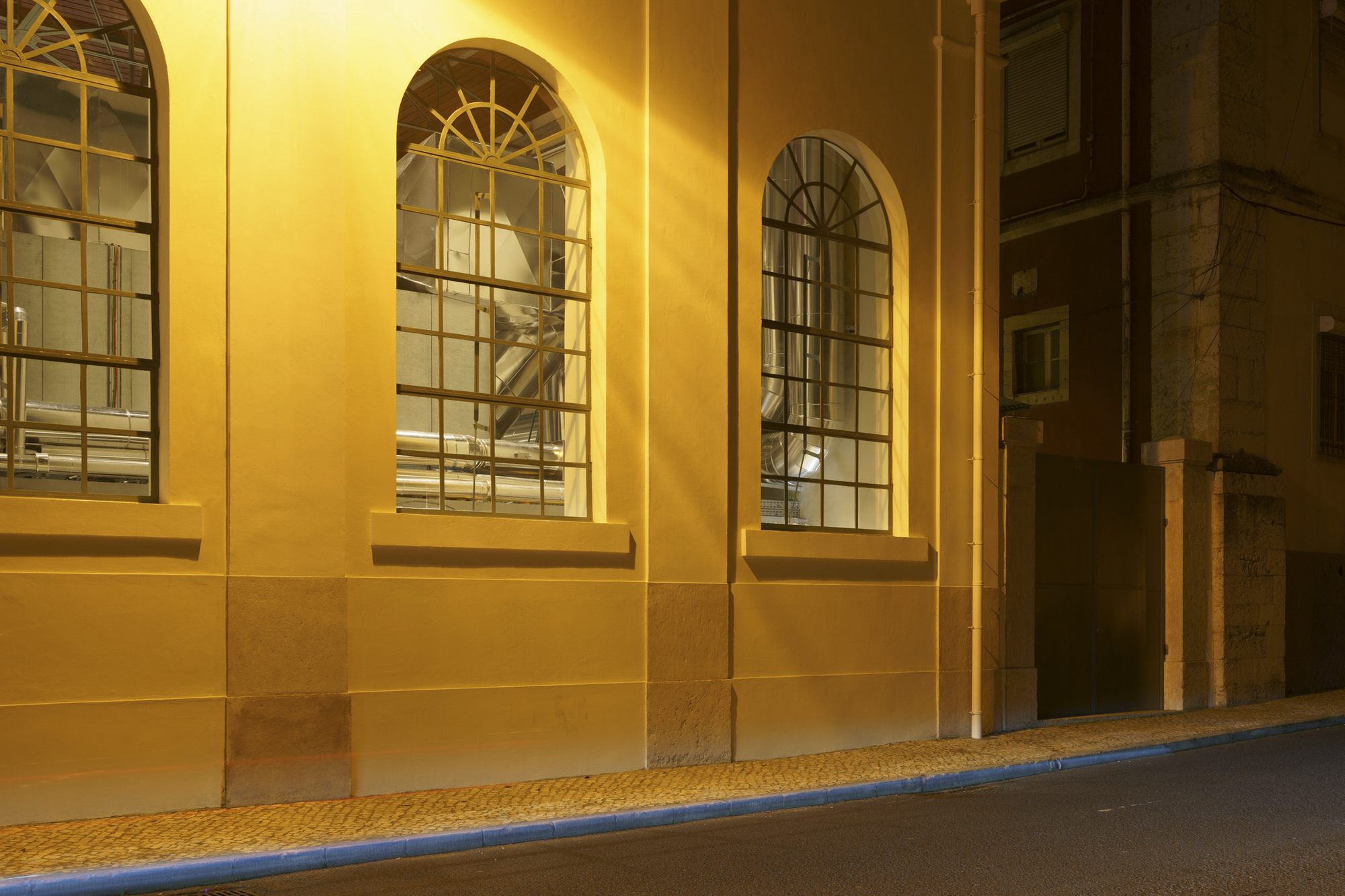 Arched windows set into the illuminated yellow-lit wall at night along the sidewalk