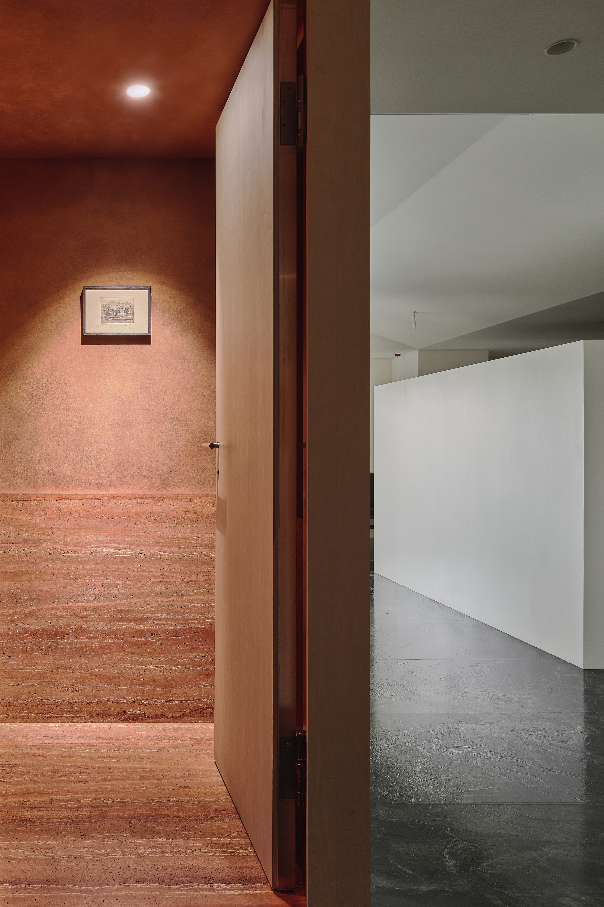 This architectural photograph showcases a doorway leading from a warm-toned, textured hallway into a minimalist, white-walled gallery space. The contrast betwee