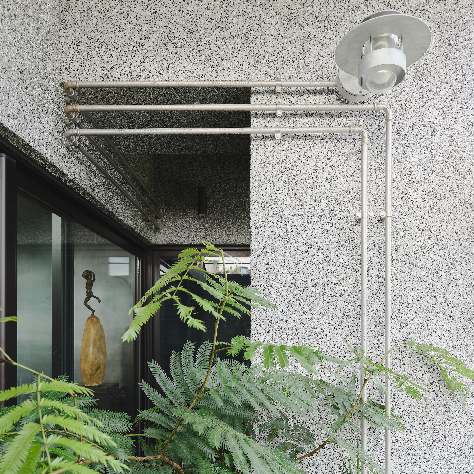 This architectural image showcases a modern building exterior with a textured terrazzo facade. A series of metal pipes and an outdoor light fixture are mounted