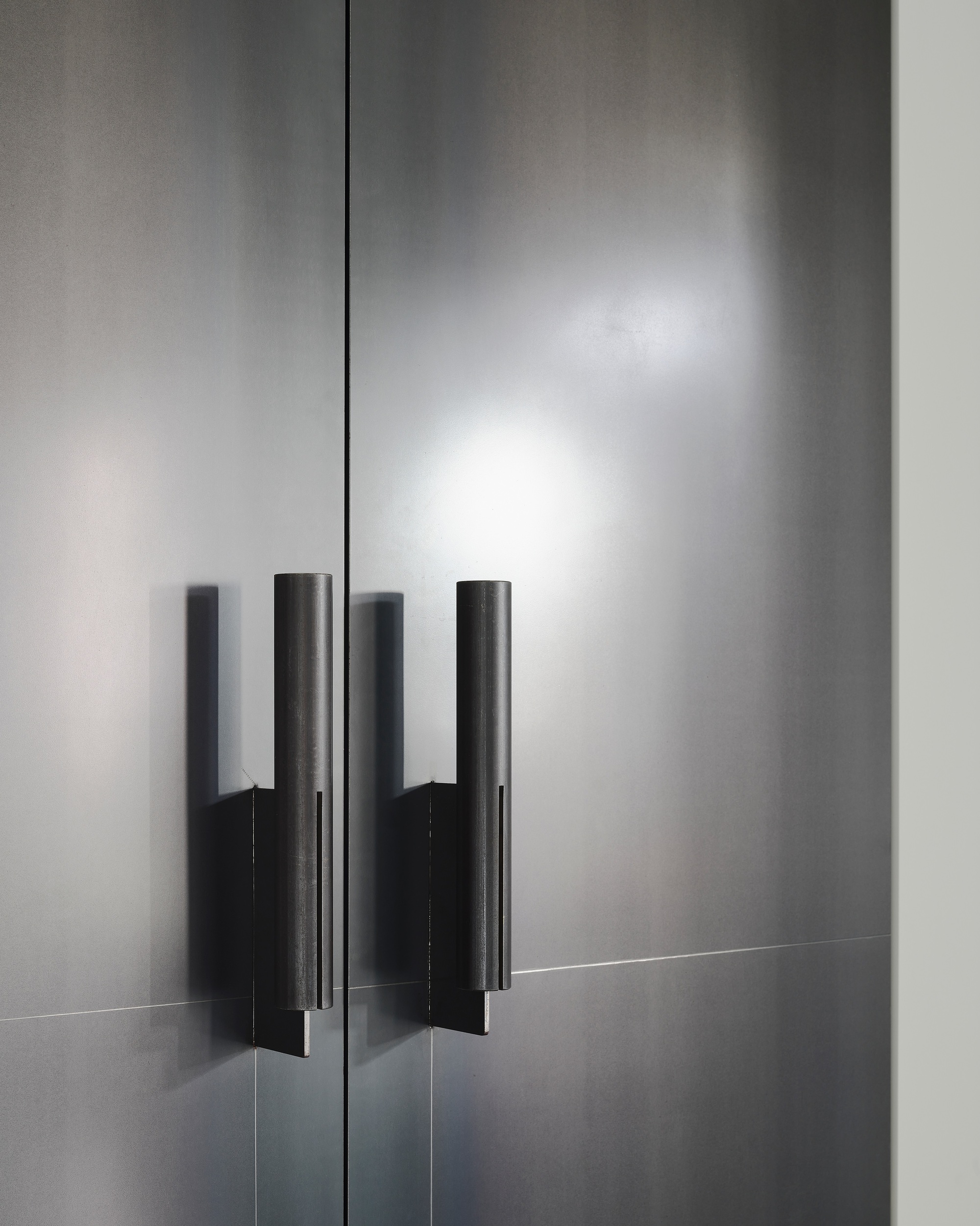 This image showcases a close-up of a modern, minimalist door with cylindrical metal handles. The gray metal surface reflects light, creating subtle gradients an