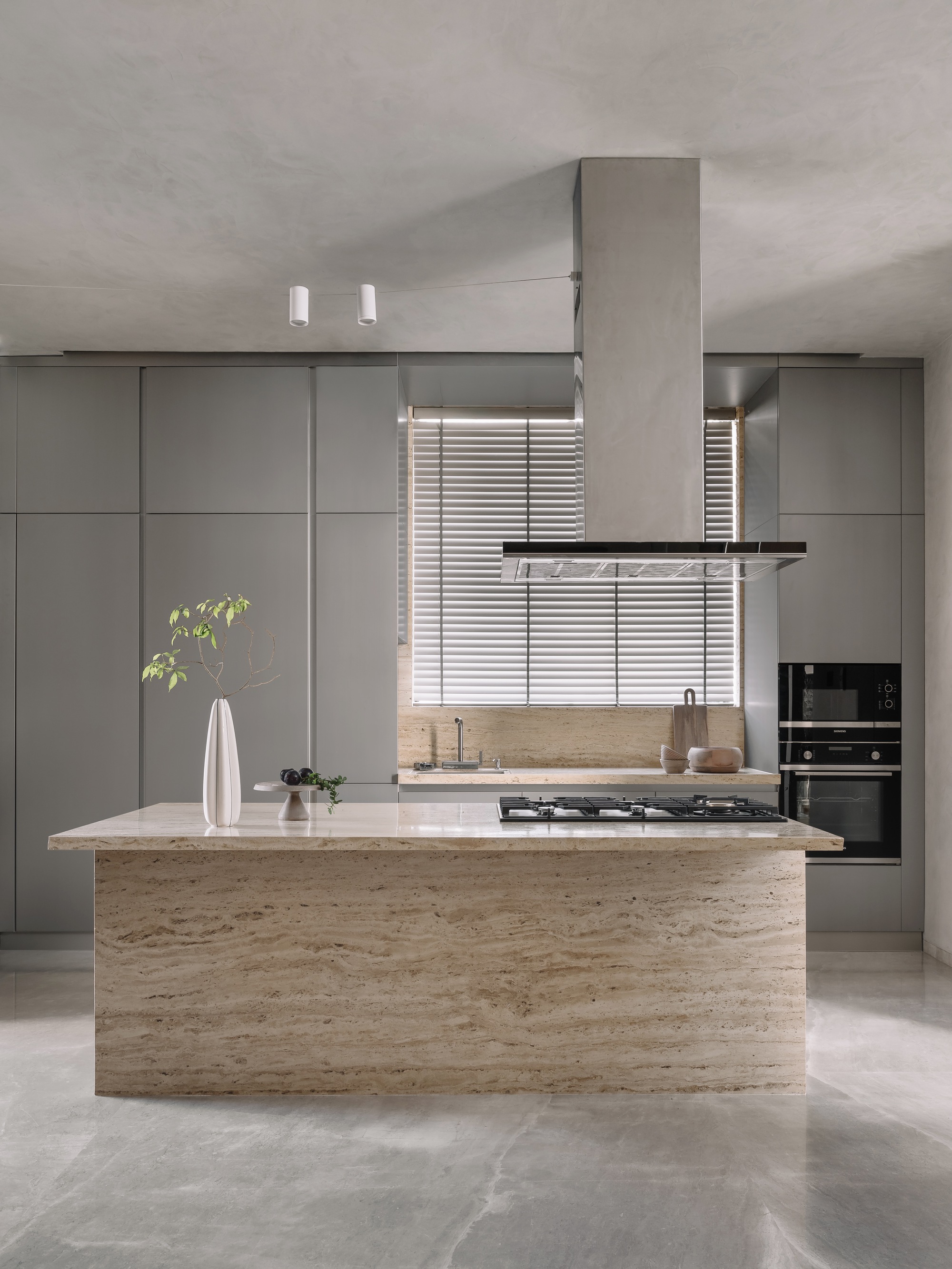 Kitchen island clad in travertine with grey cabinetry and timber blinds filtering window light