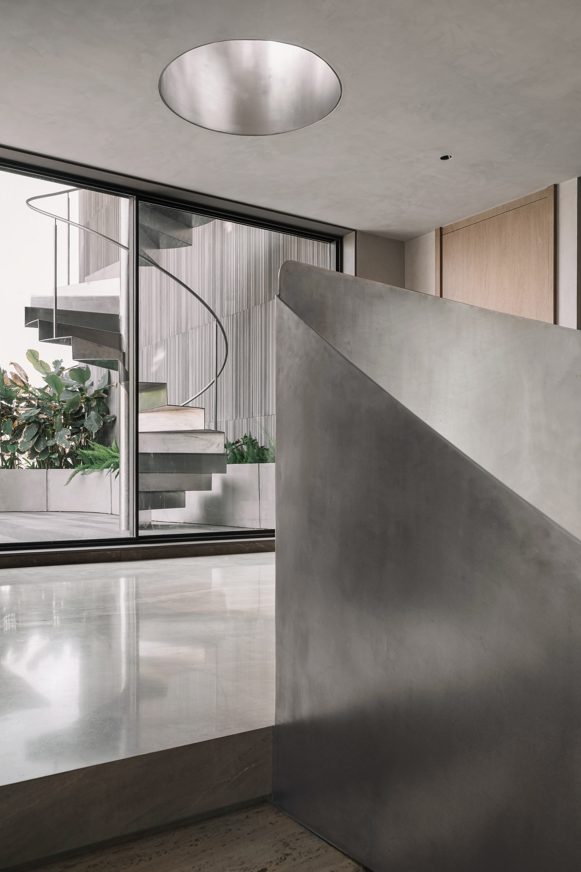 Curved polished concrete wall beside glazed opening revealing spiral stair and planted courtyard beyond