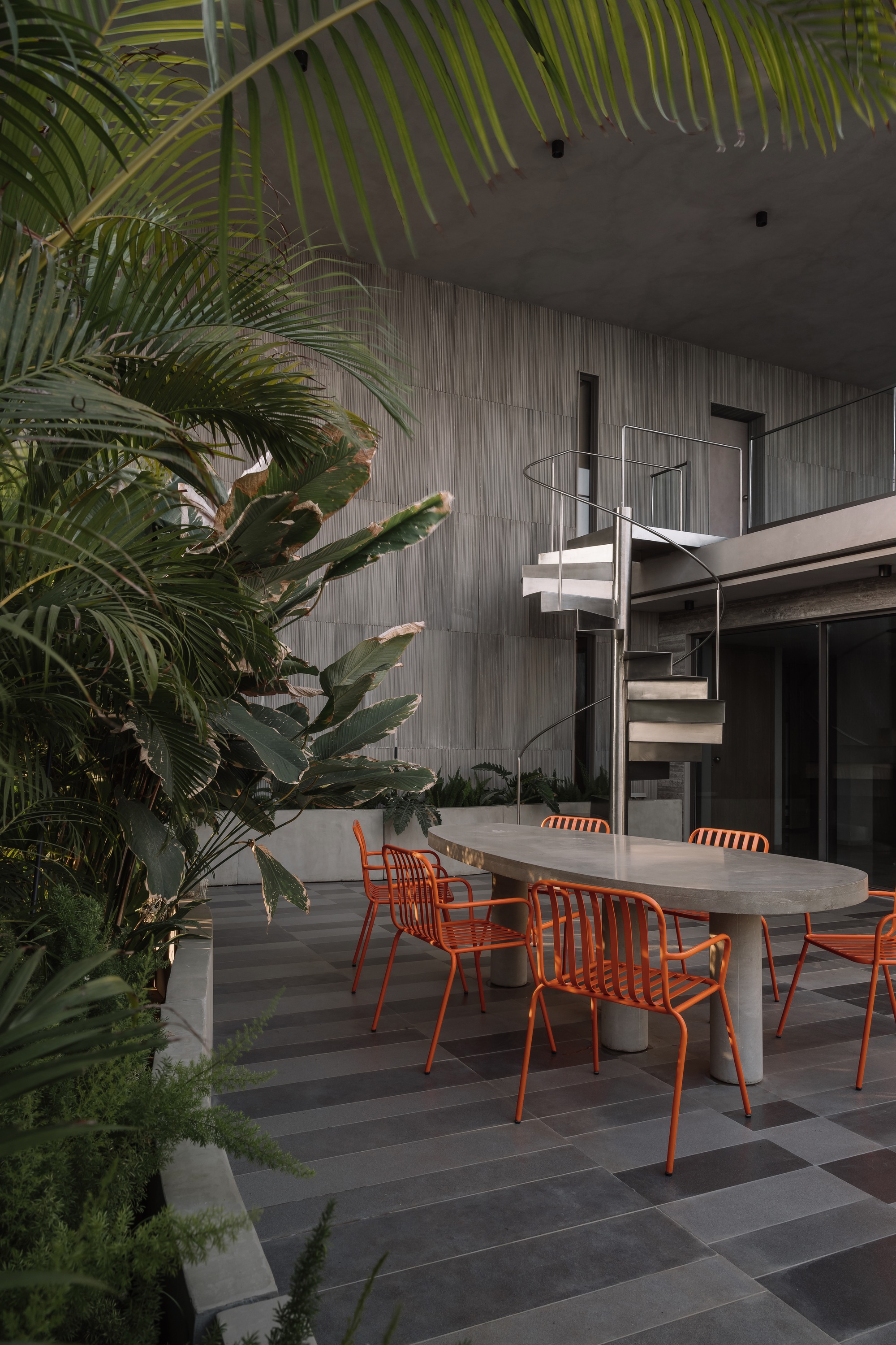 Outdoor dining terrace with concrete table and orange metal chairs surrounded by tropical palms at dusk
