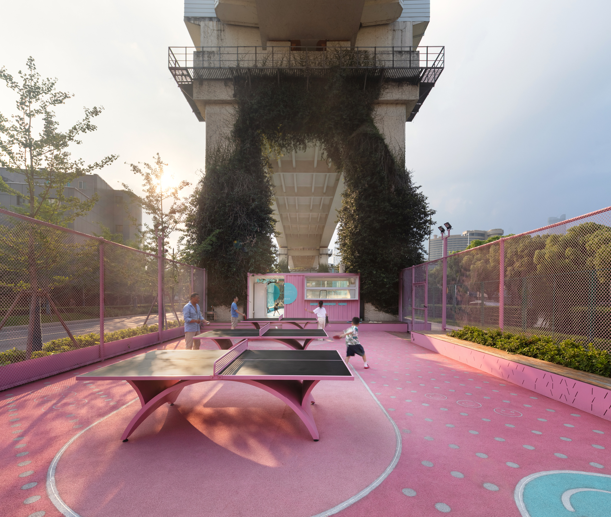 Pink recreation court with ping-pong tables beneath a concrete viaduct with vegetation-clad piers at dusk