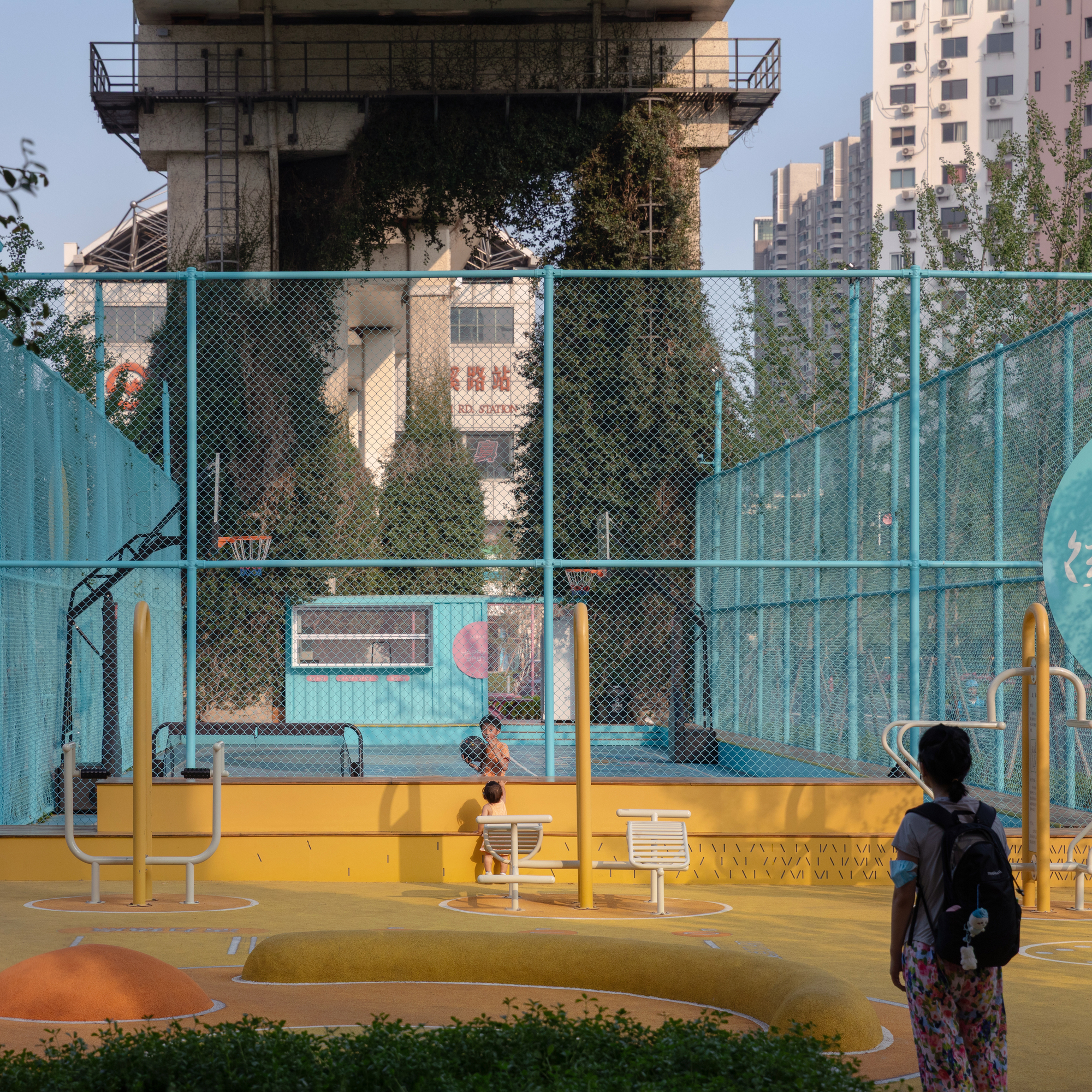 Outdoor basketball court with yellow base and turquoise mesh fencing beneath a concrete elevated structure