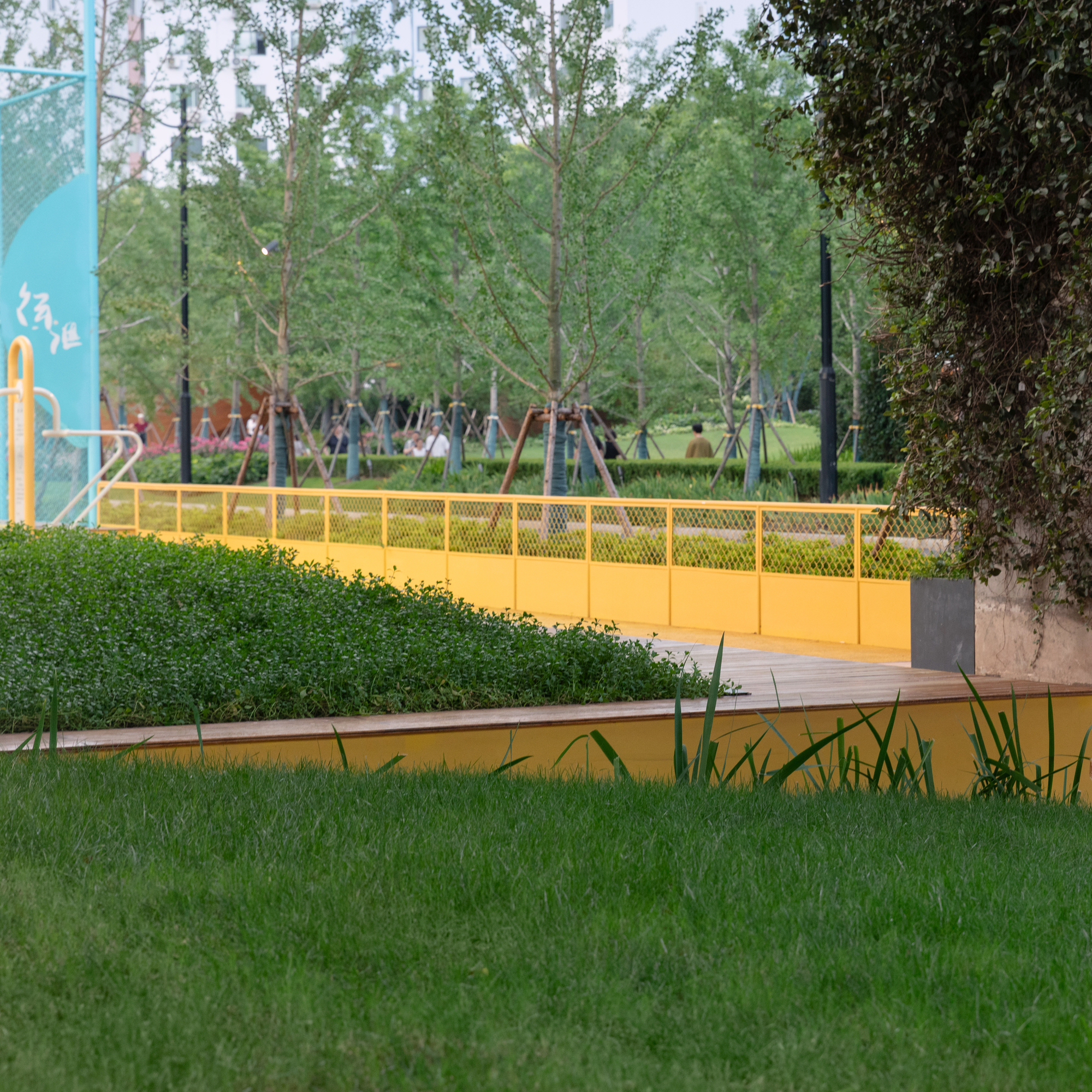 Yellow mesh bridge crossing a water feature with trimmed hedge and lawn beneath established trees