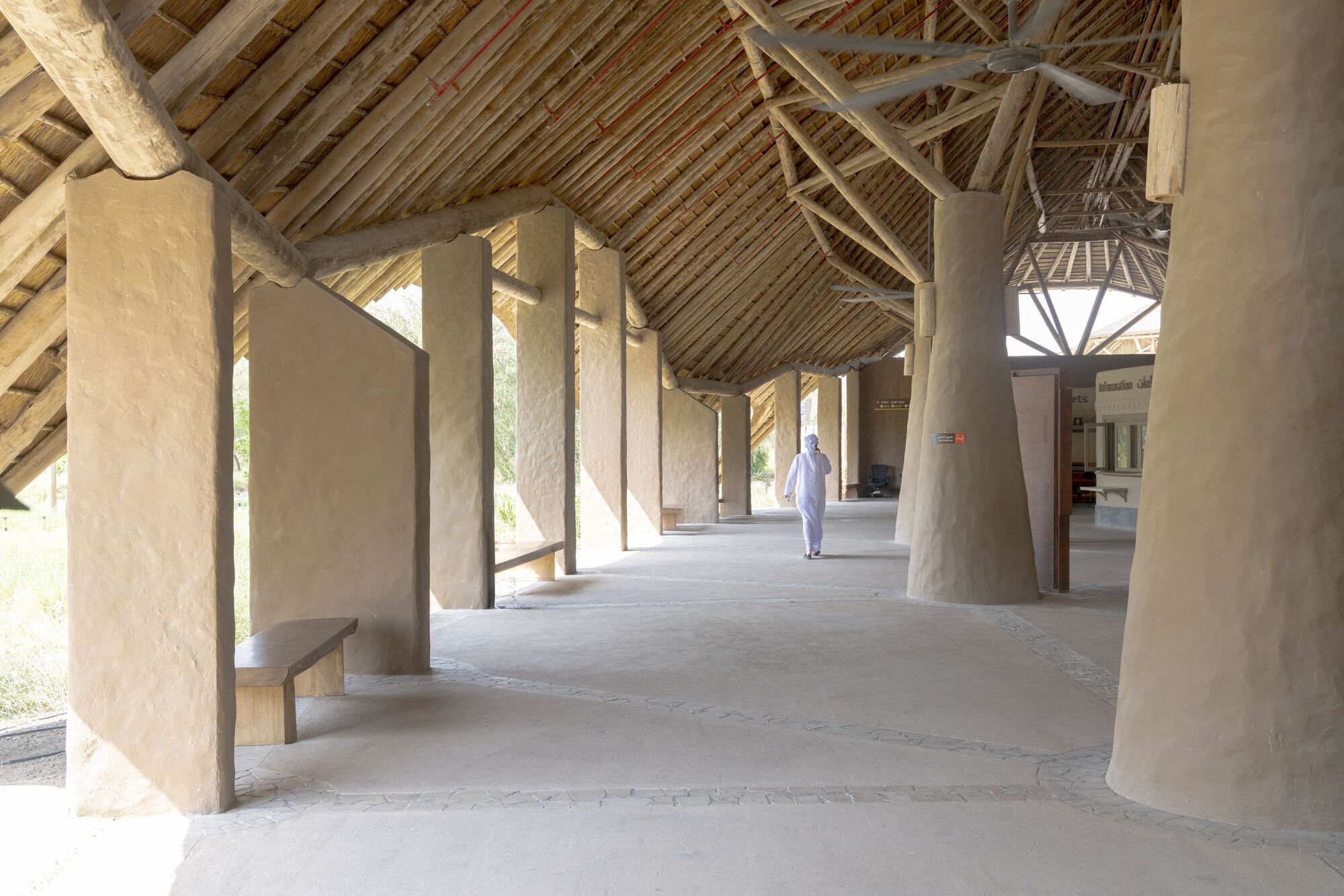 Covered walkway with plastered earthen walls and exposed timber roof structure as figure walks through