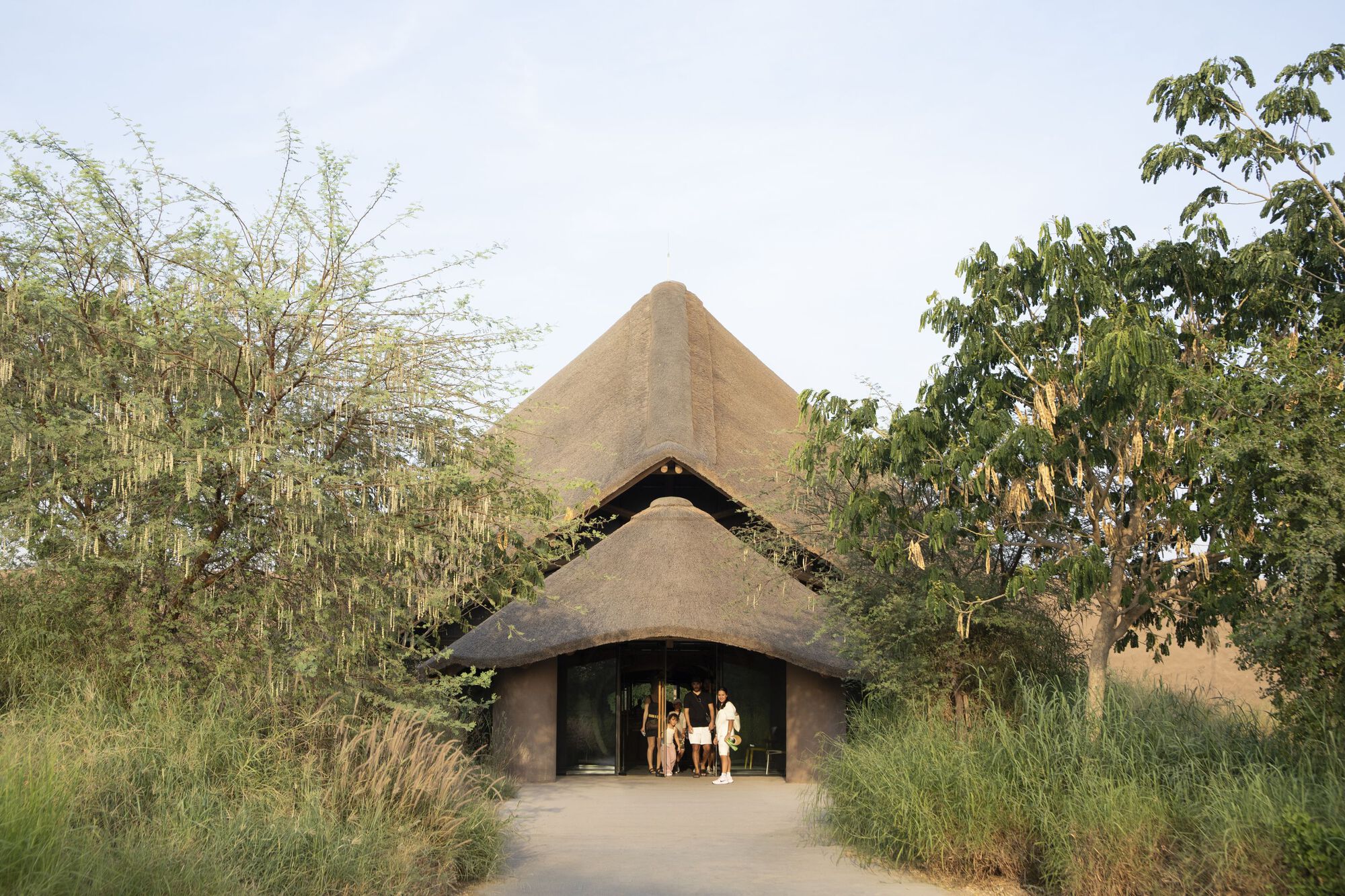 Entry pavilion with layered thatched roofs and arched opening as visitors gather at threshold