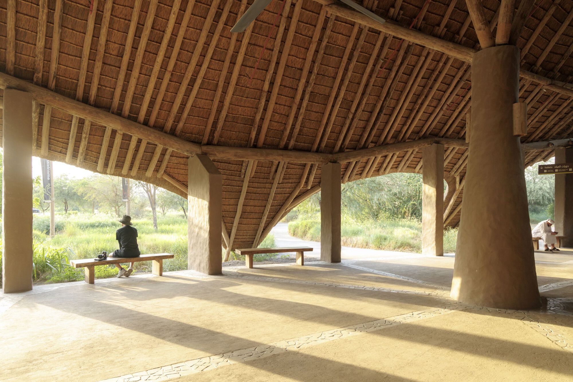 Open-air pavilion interior with exposed timber rafters and thatched ceiling sheltering benches and a seated figure