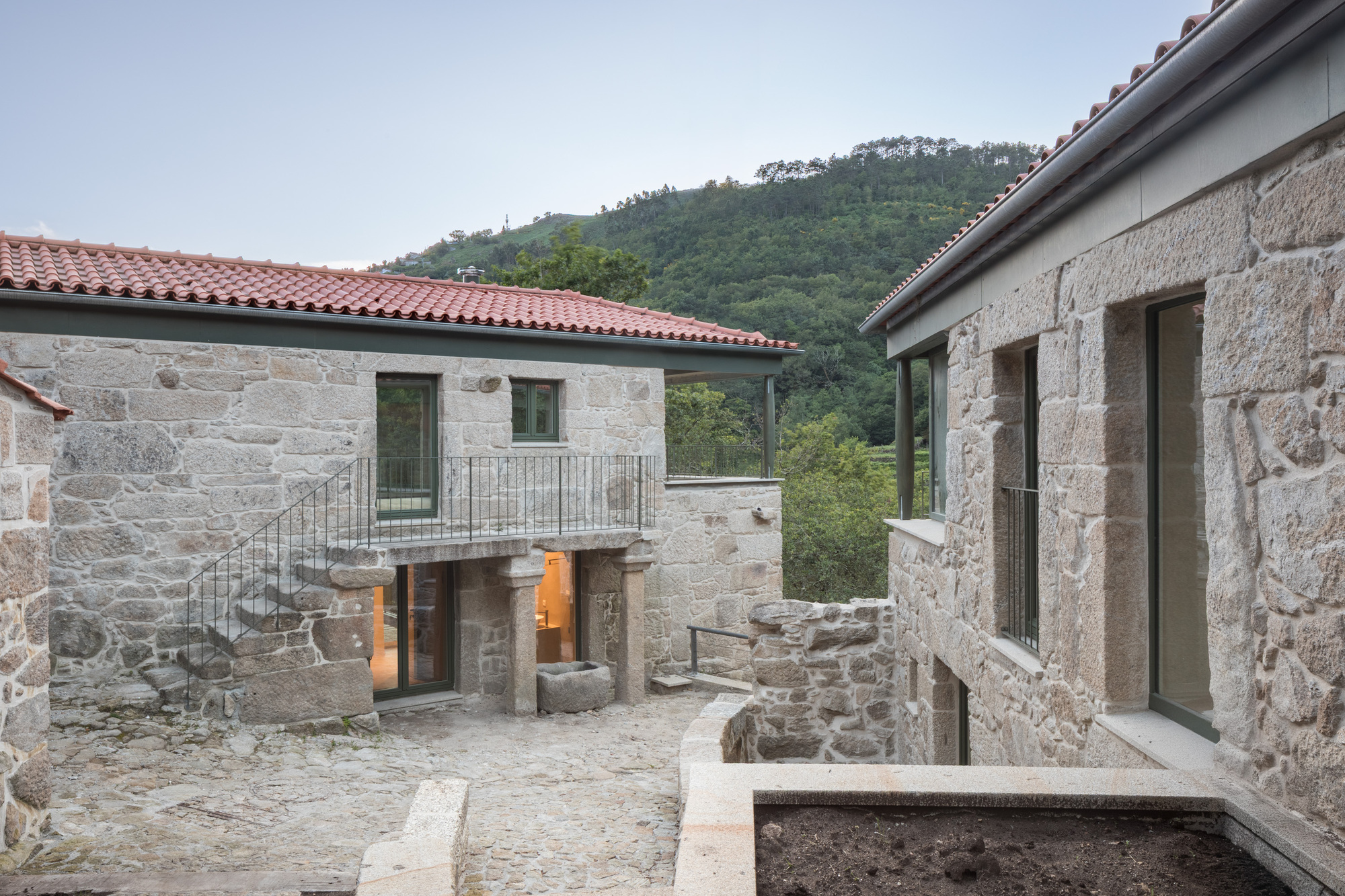 Stone courtyard enclosed by restored masonry buildings with terracotta tile roofs against a forested hillside