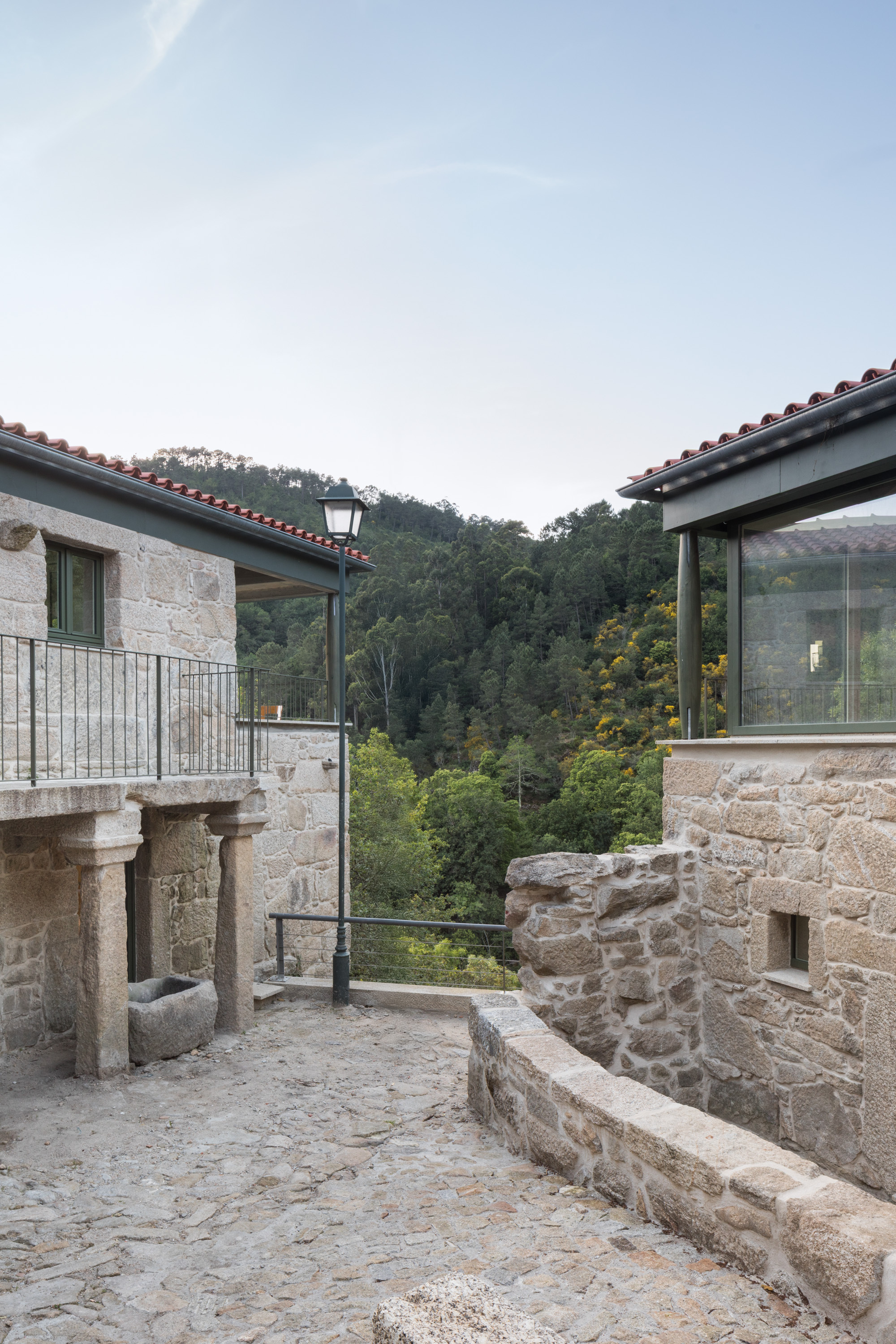 Stone courtyard terrace with historic column fragments and contemporary glazing frames opening to the wooded hillside at dusk