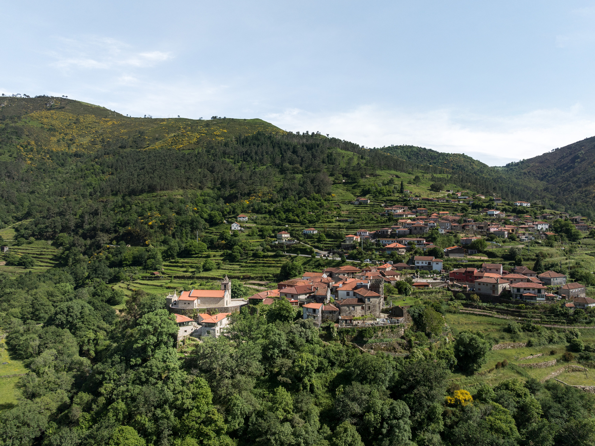 Hillside village with terracotta roofed houses dispersed across agricultural terraces between forested slopes under afternoon light