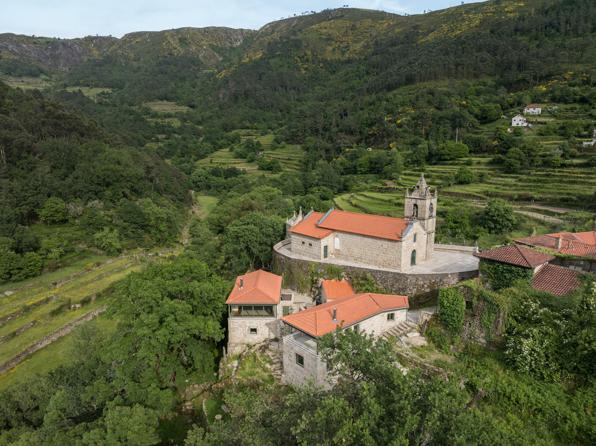 Aerial view of stone buildings with terracotta roofs nestled among terraced fields and forested hillsides